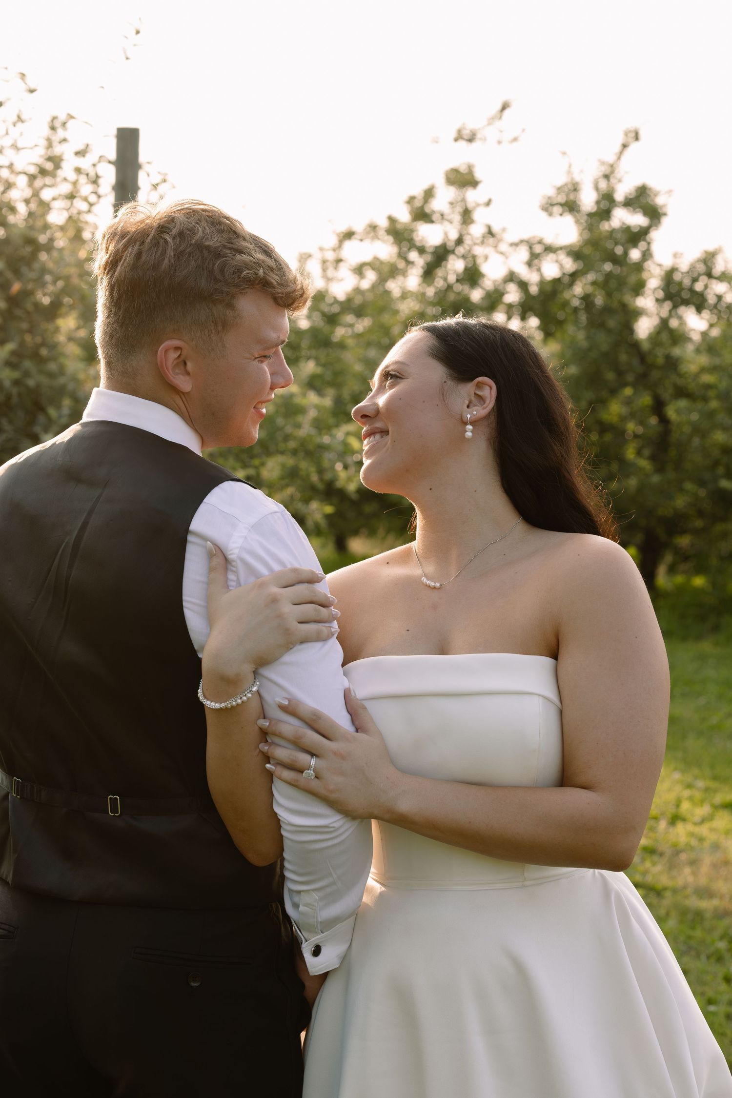 Bride and groom share a romantic moment in a sunlit orchard, smiling at each other in wedding attire.