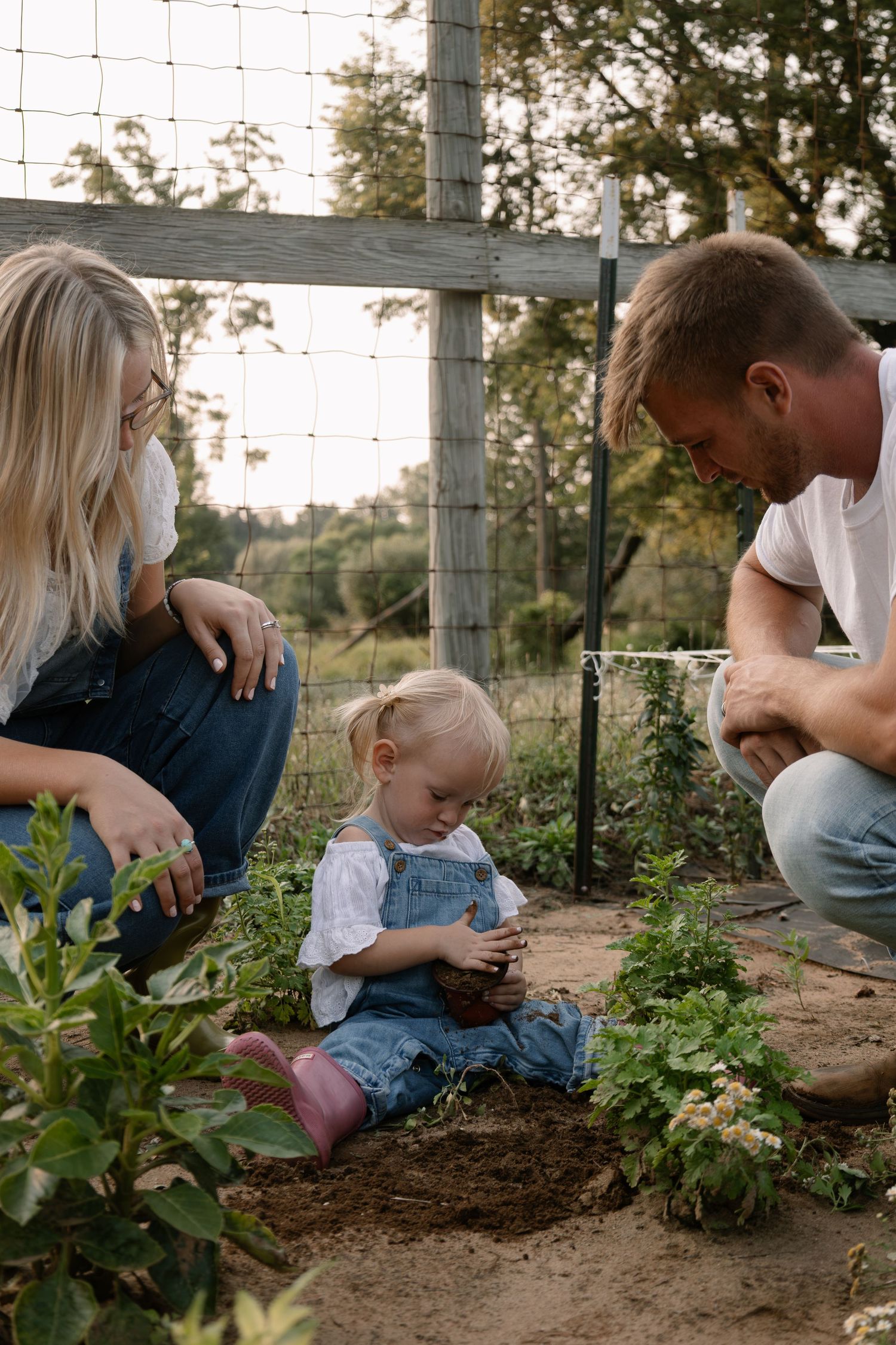 Parents watch their toddler daughter play with soil in a garden bed near a wooden fence during a family outdoor activity.