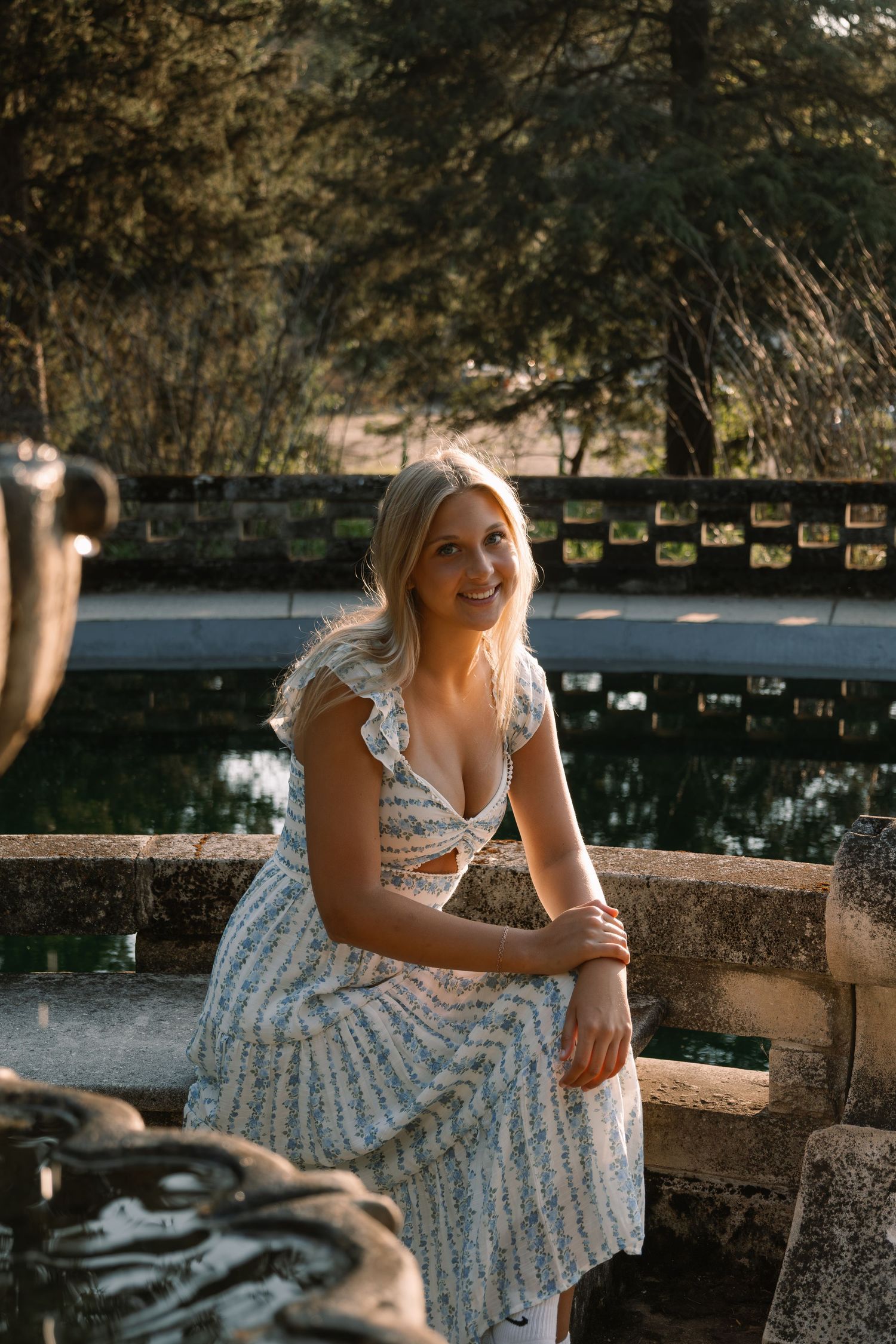 Smiling blonde woman in floral dress sitting on stone wall by reflective pond in sunlit garden.