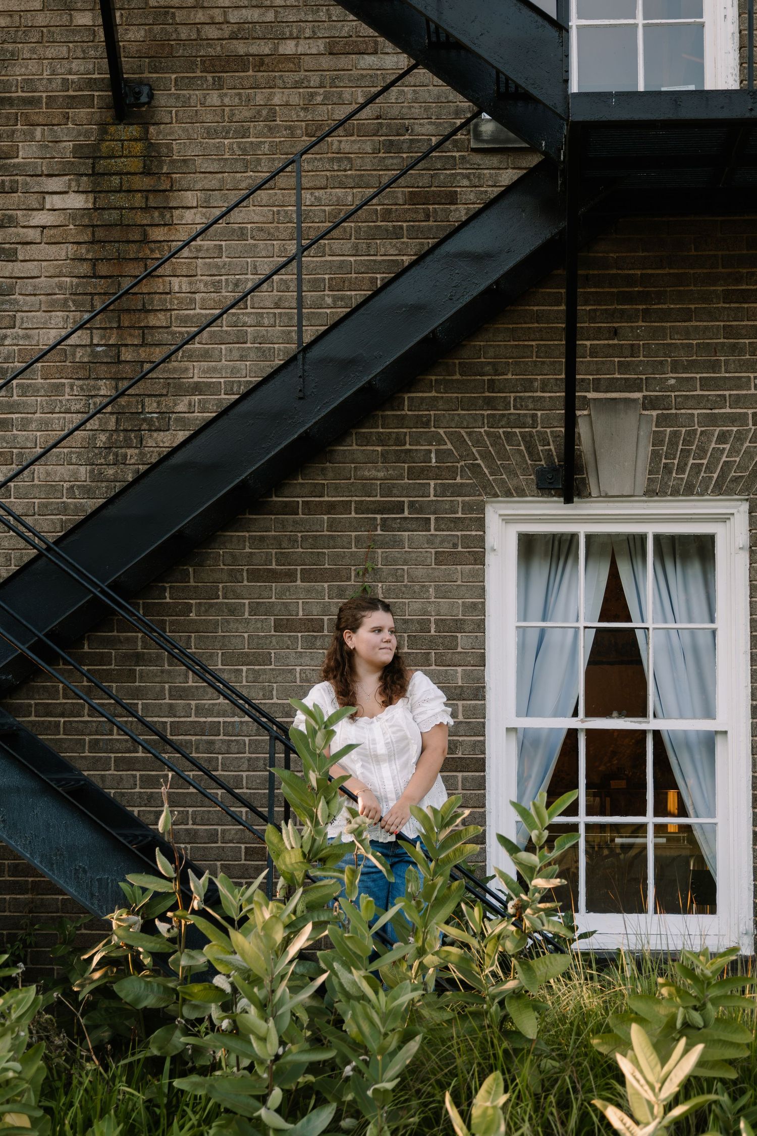 Young woman in white top and jeans stands among green plants beside brick building with black metal fire escape stairs.