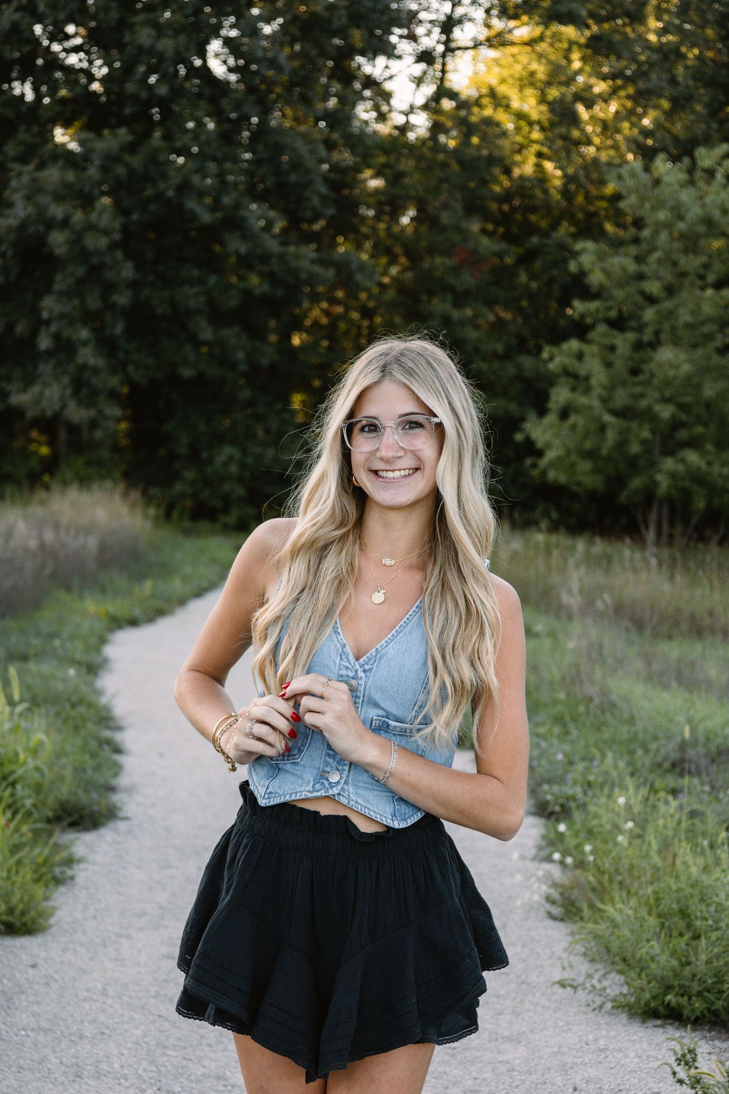 Smiling blonde woman in glasses, denim crop top and black skirt standing on a gravel path surrounded by trees.