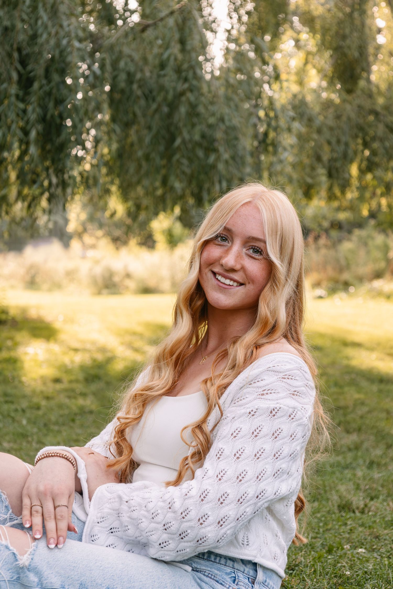 Young blonde woman smiling in white knit cardigan and jeans, sitting on grass under a willow tree in golden sunlight.