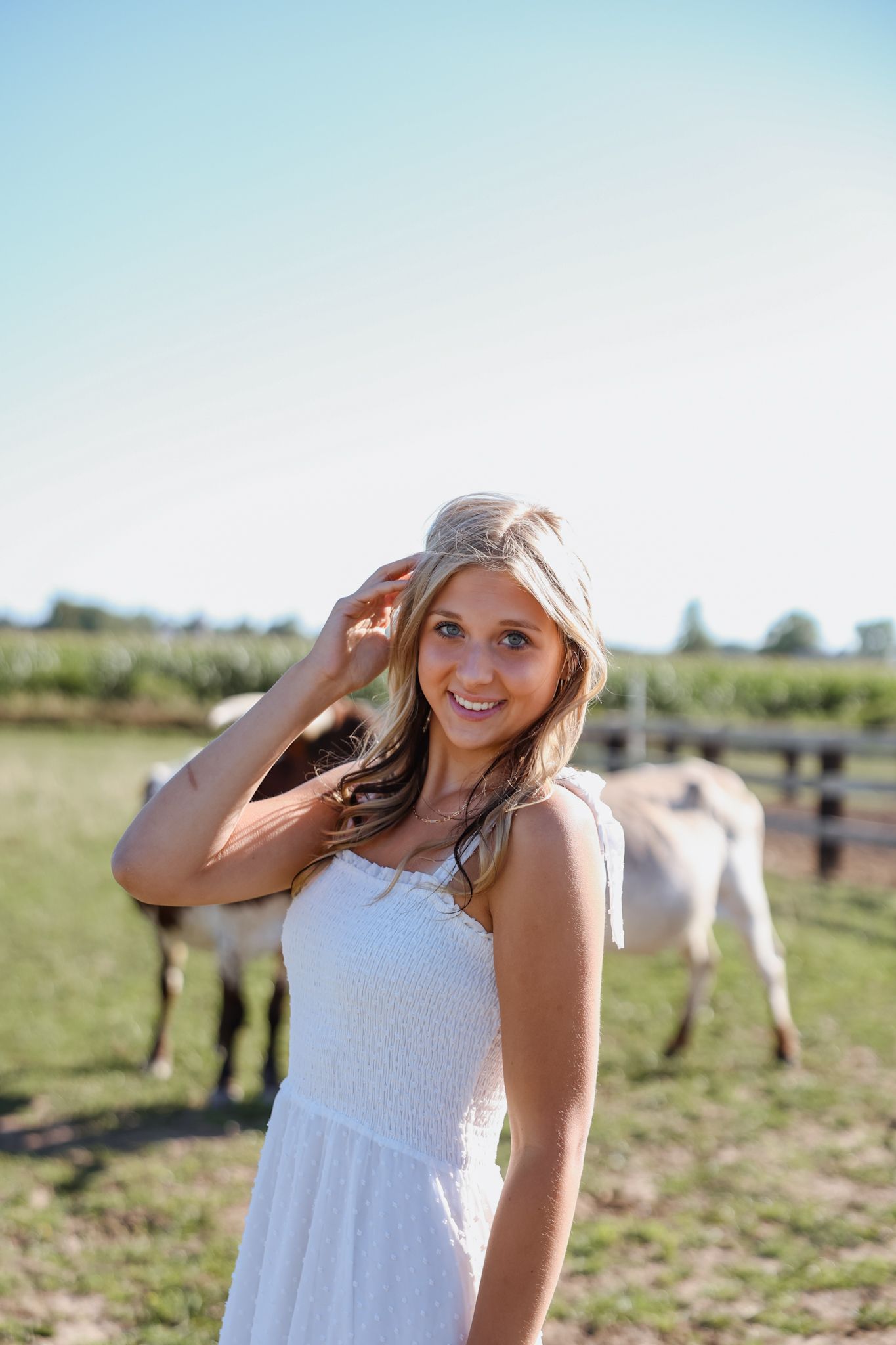 Blonde woman in white sundress smiling outdoors near horses in a sunny farm field setting.