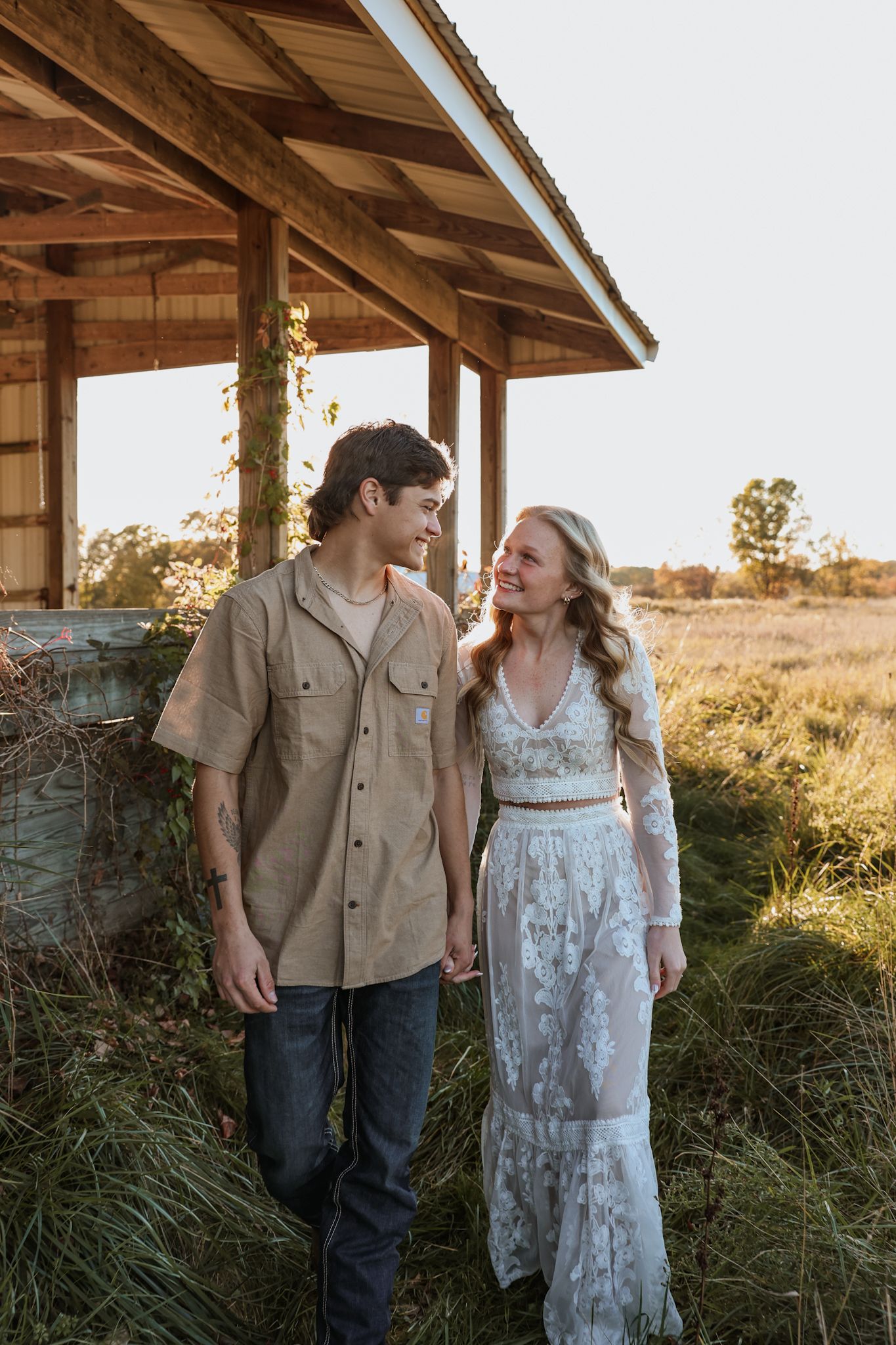 Smiling engaged couple holding hands and walking near a rustic wooden structure in a golden field.