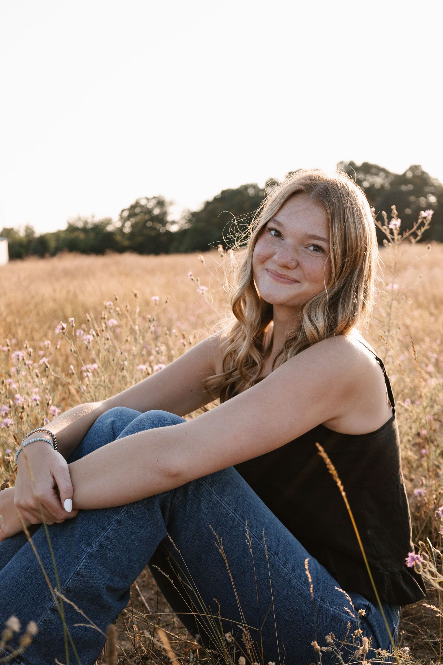 Young woman in black tank top and jeans sitting in a golden field of wildflowers at sunset.