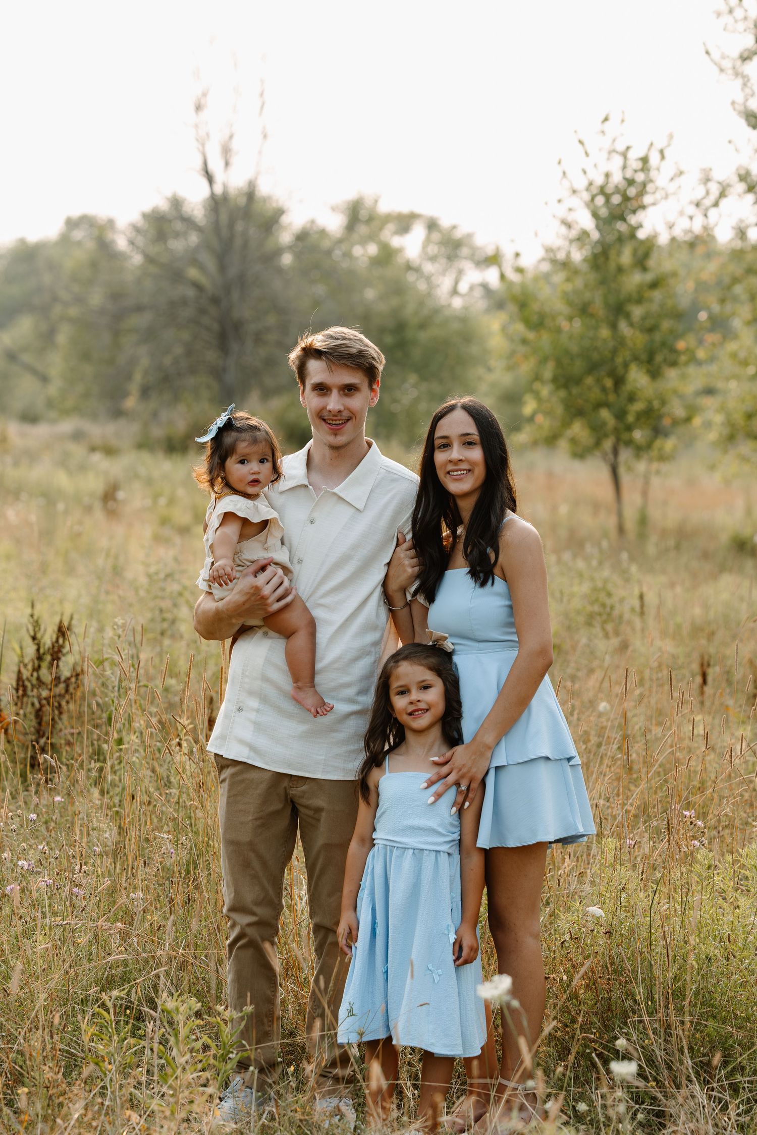 Young family of four posing outdoors in a sunny meadow, parents with two daughters in matching blue outfits.