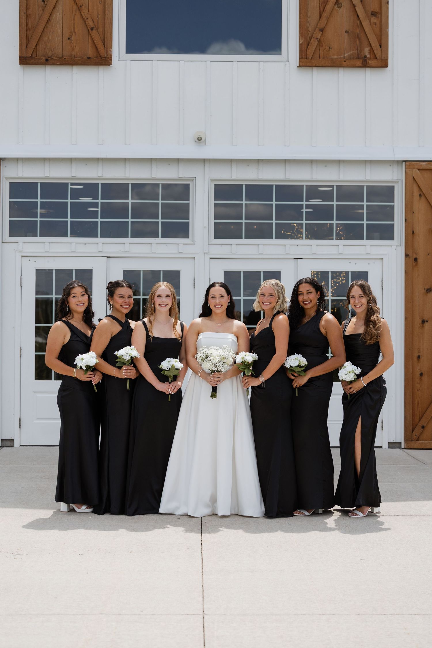 Bride in white gown poses with six bridesmaids in black dresses holding white bouquets outside a white barn.