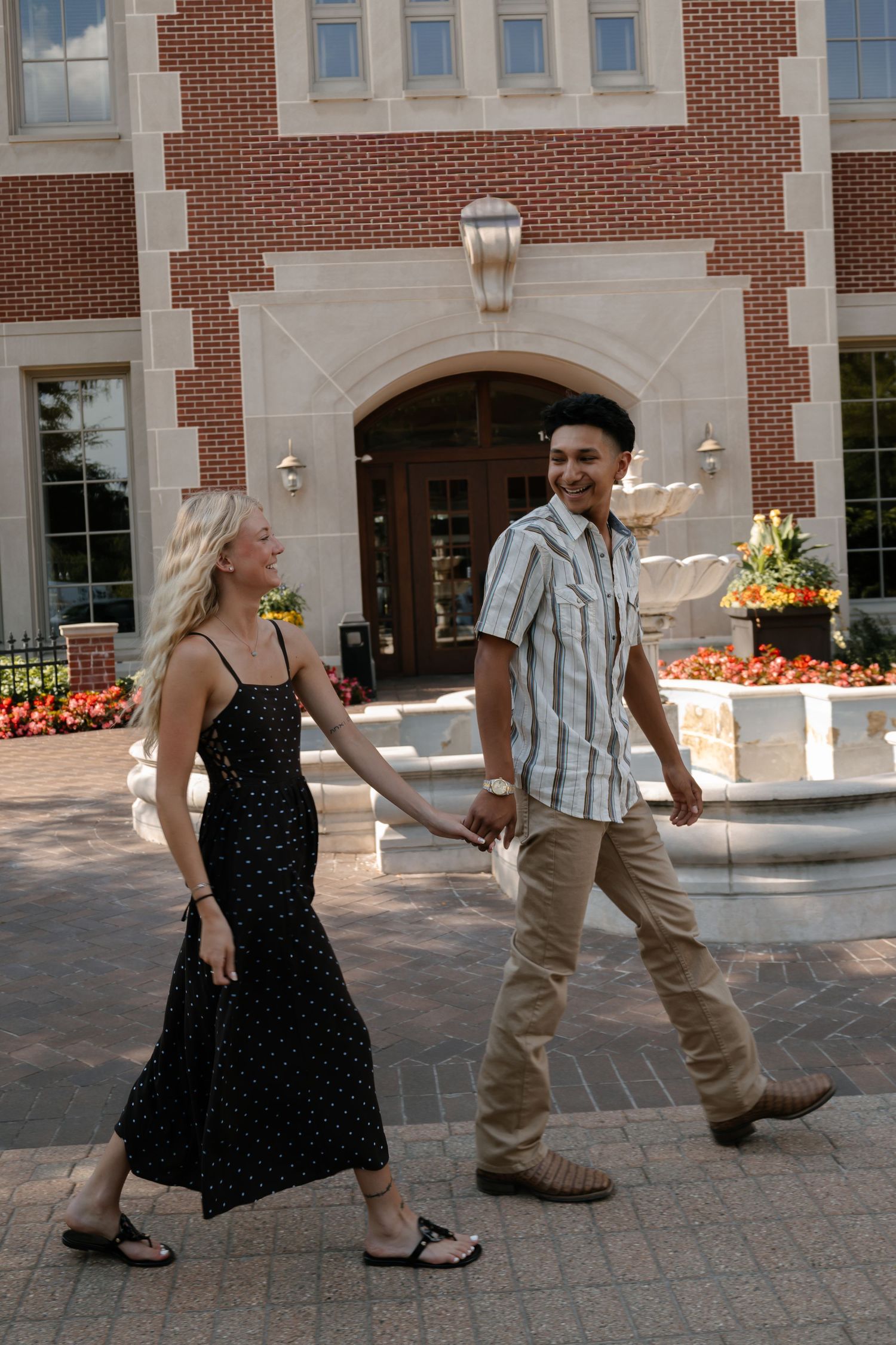 A smiling couple holds hands and walks together outside a brick building with flower gardens on a sunny day.