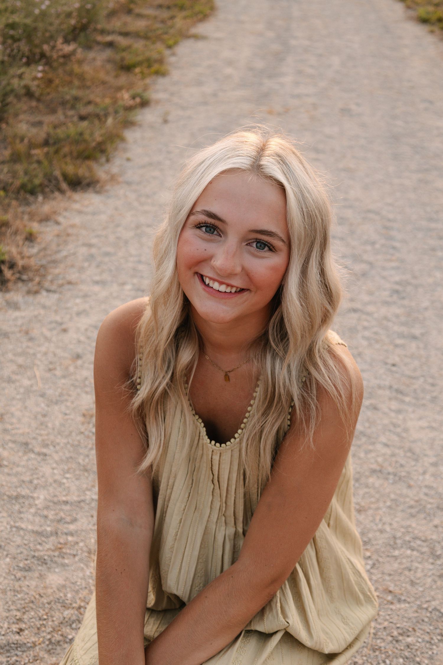 Smiling young blonde woman in a light olive dress sitting on a dirt path outdoors in warm sunlight.