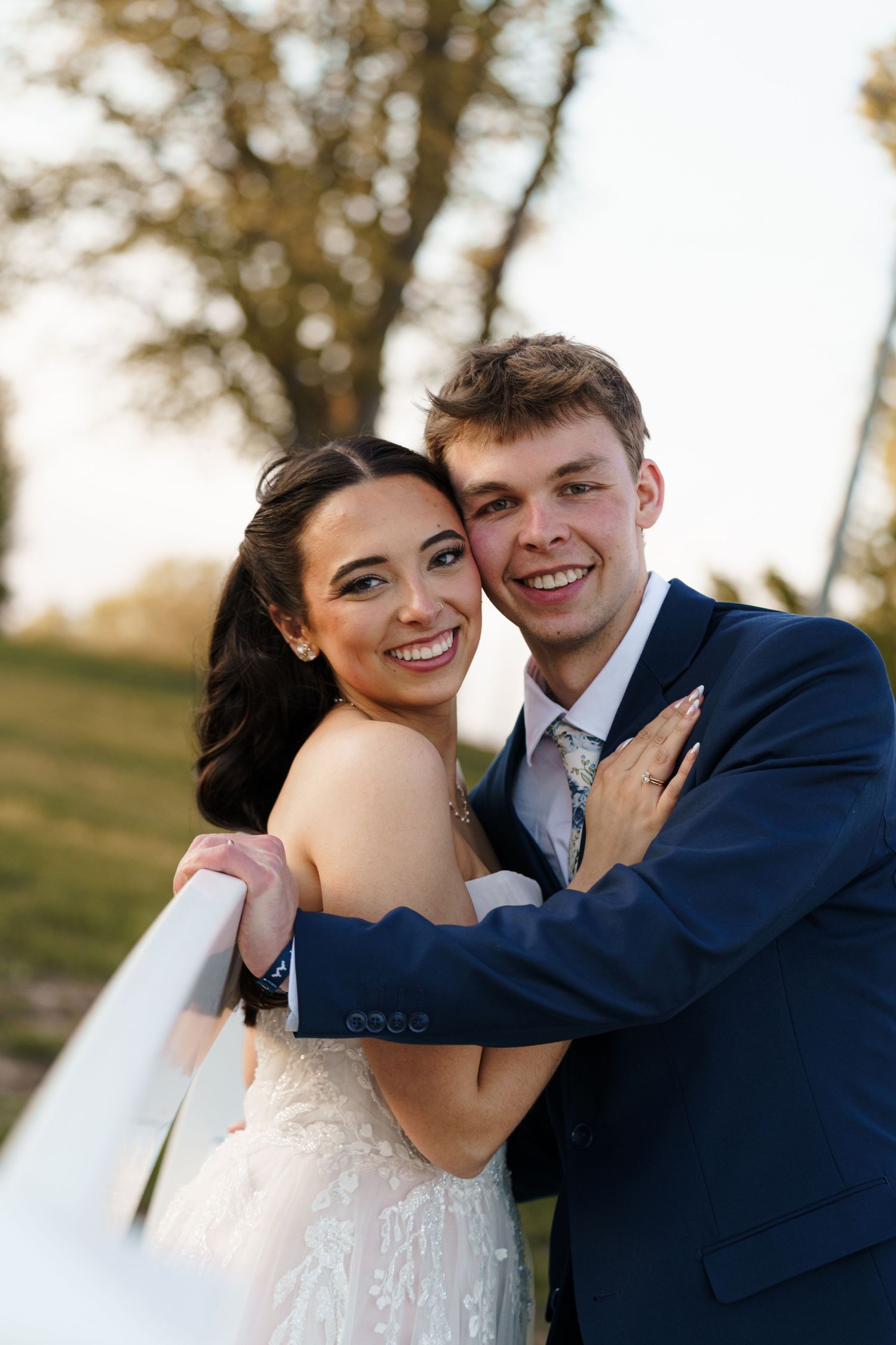 Joyful bride and groom embrace near a white fence at golden hour during their wedding photo session.