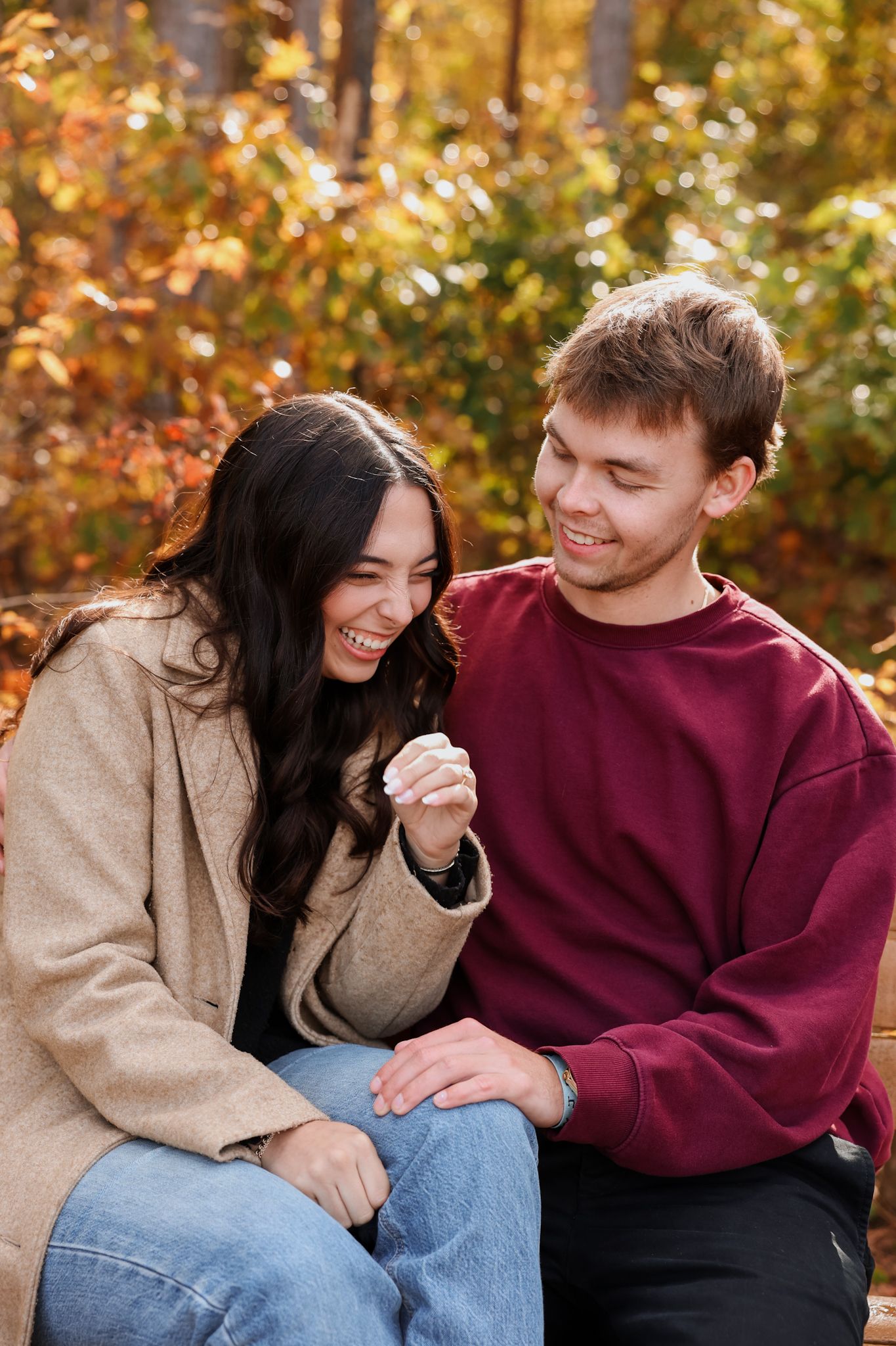 A laughing couple sits together on a bench in an autumn park, holding hands and sharing a joyful moment.