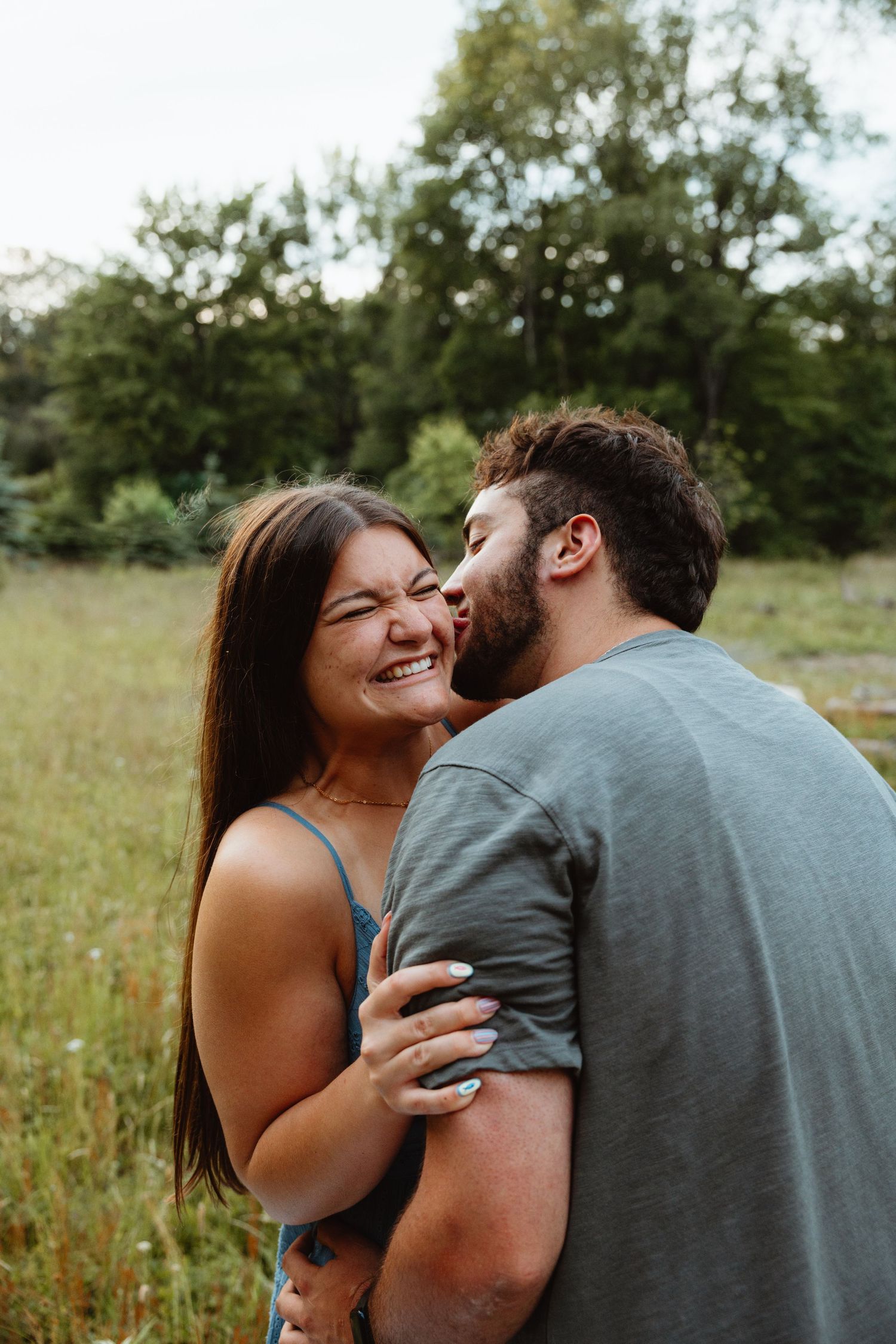Man kissing woman on the cheek as she laughs joyfully in a lush green outdoor field setting.