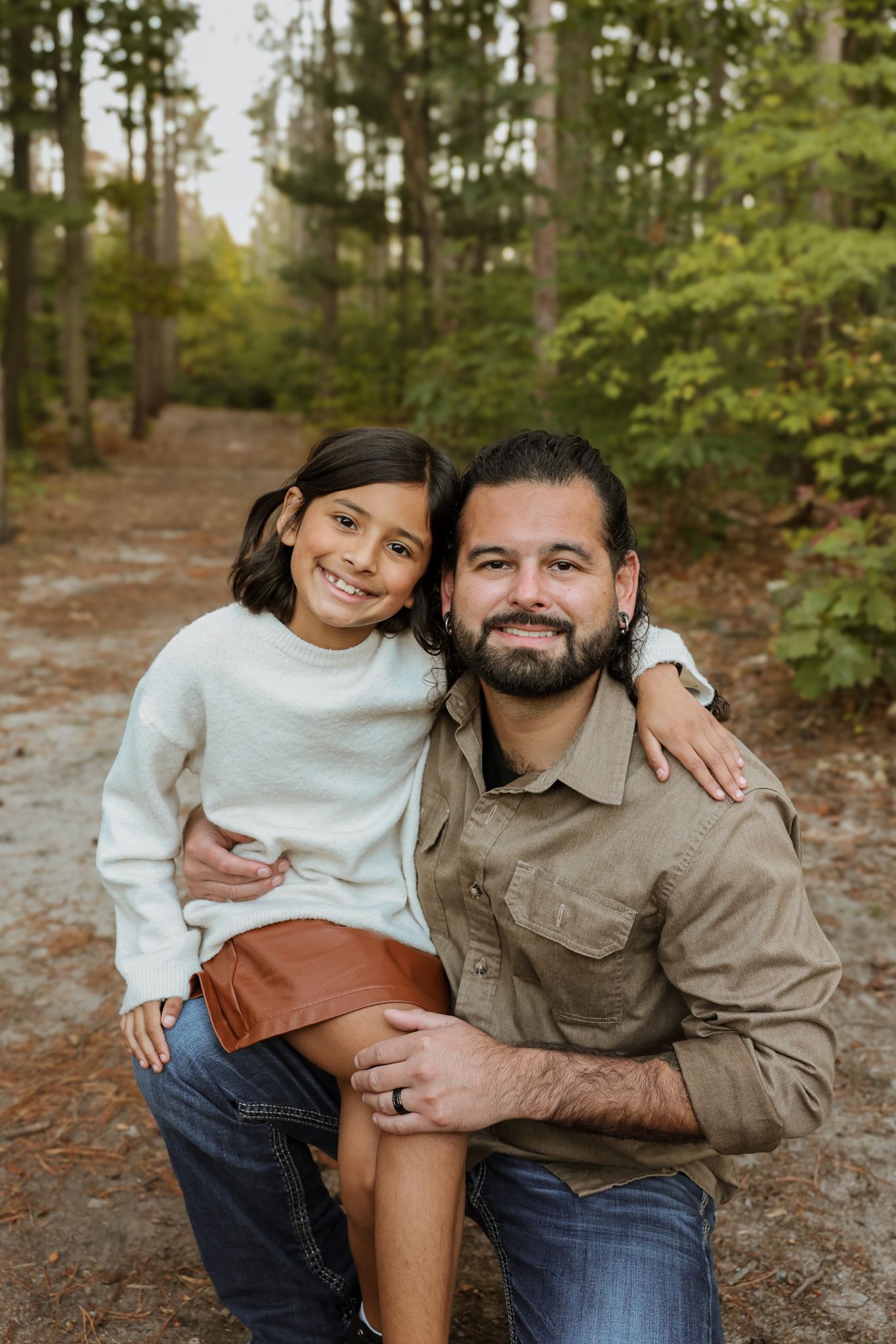 Father kneels beside smiling daughter in white sweater during outdoor forest photo session.