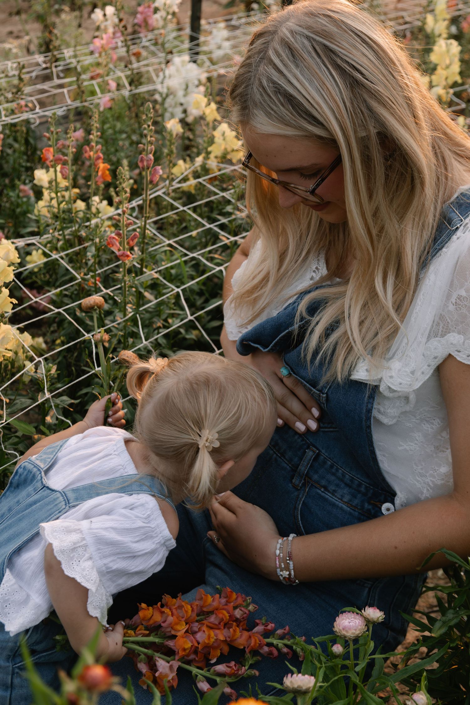 Blonde mother in denim overalls and glasses tends to colorful garden flowers with young toddler daughter.
