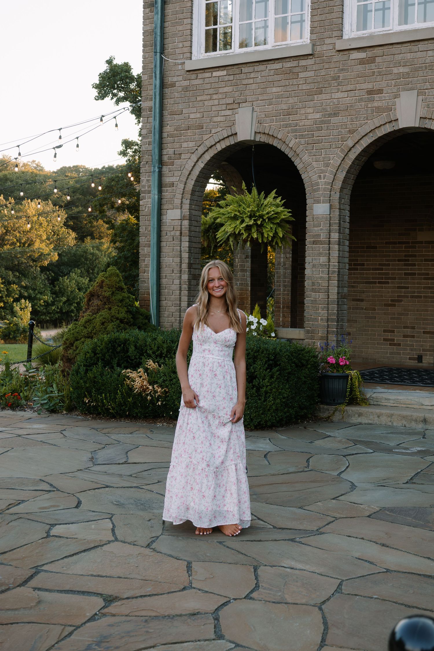 Woman in floral maxi dress standing on stone patio outside elegant brick building with arched doorways.