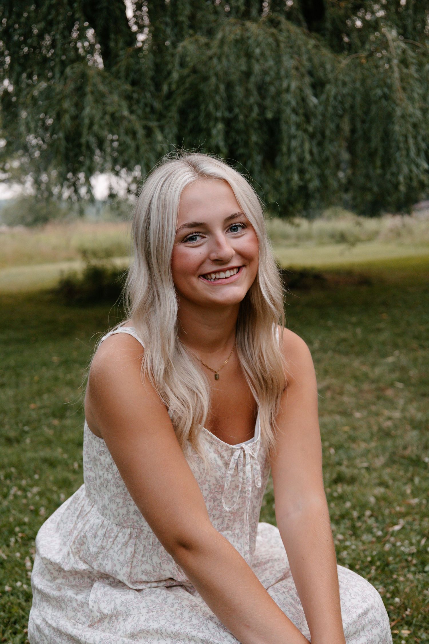 Smiling blonde woman in a floral sundress sitting outdoors on grass near a weeping willow tree.