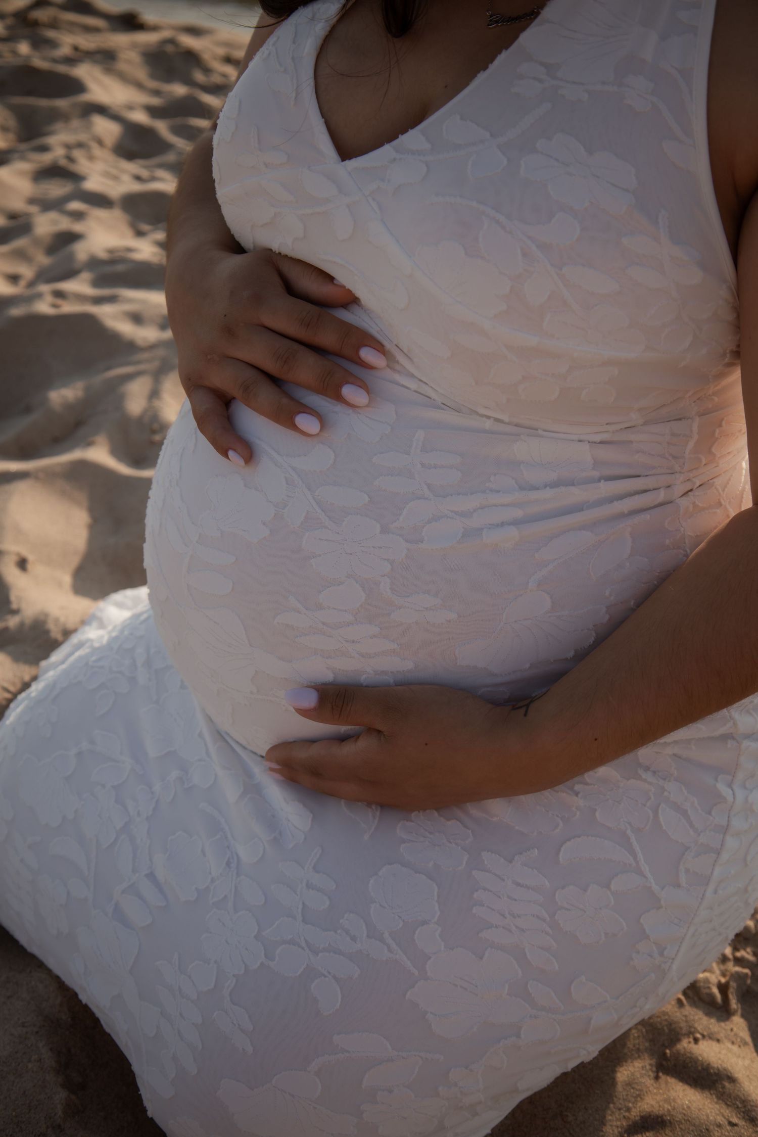 Pregnant woman in white lace dress cradling baby bump on sandy beach.