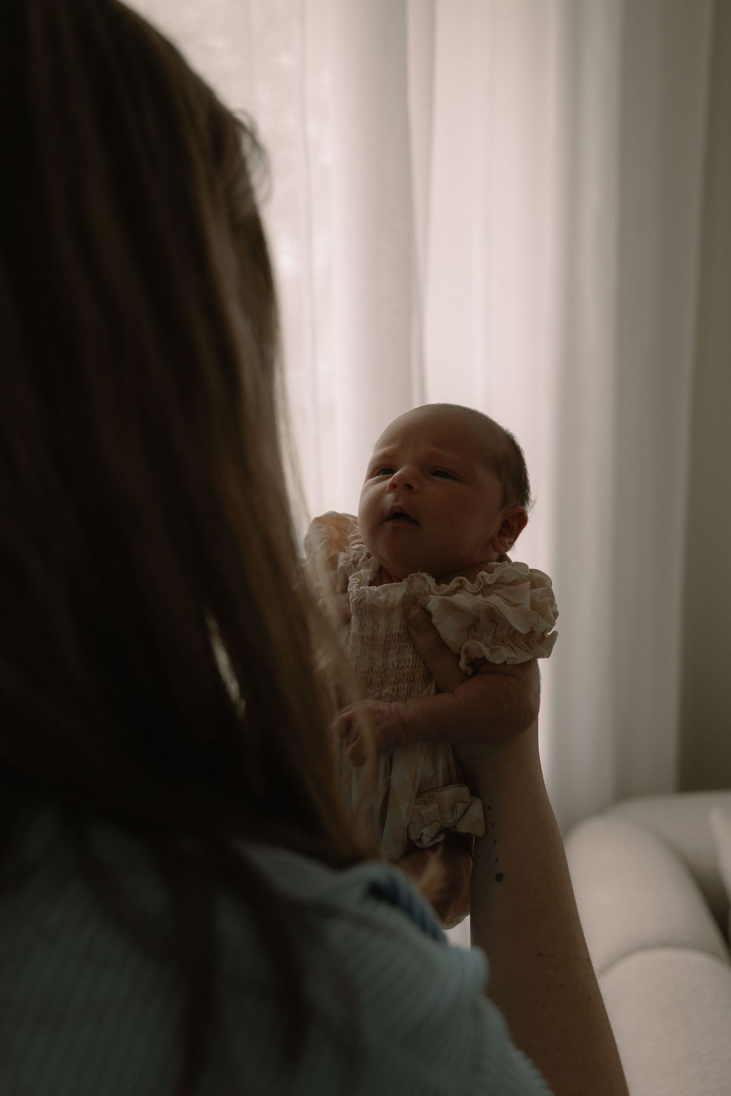 Mother gently holding her newborn baby up near a bright window in a softly lit nursery room.
