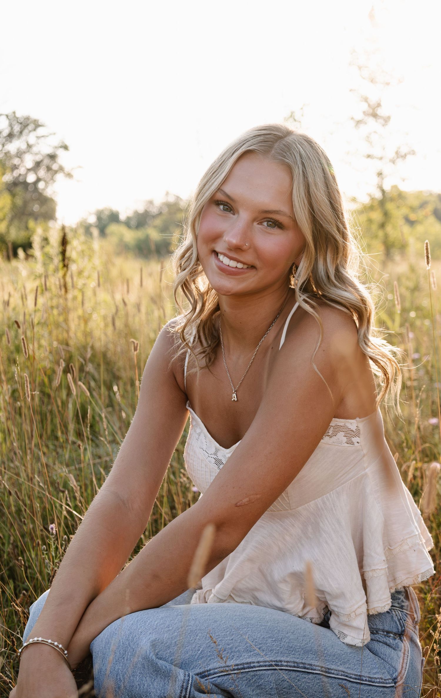 Young blonde woman smiling in a sunlit field, wearing a white top and jeans, golden hour outdoor portrait.