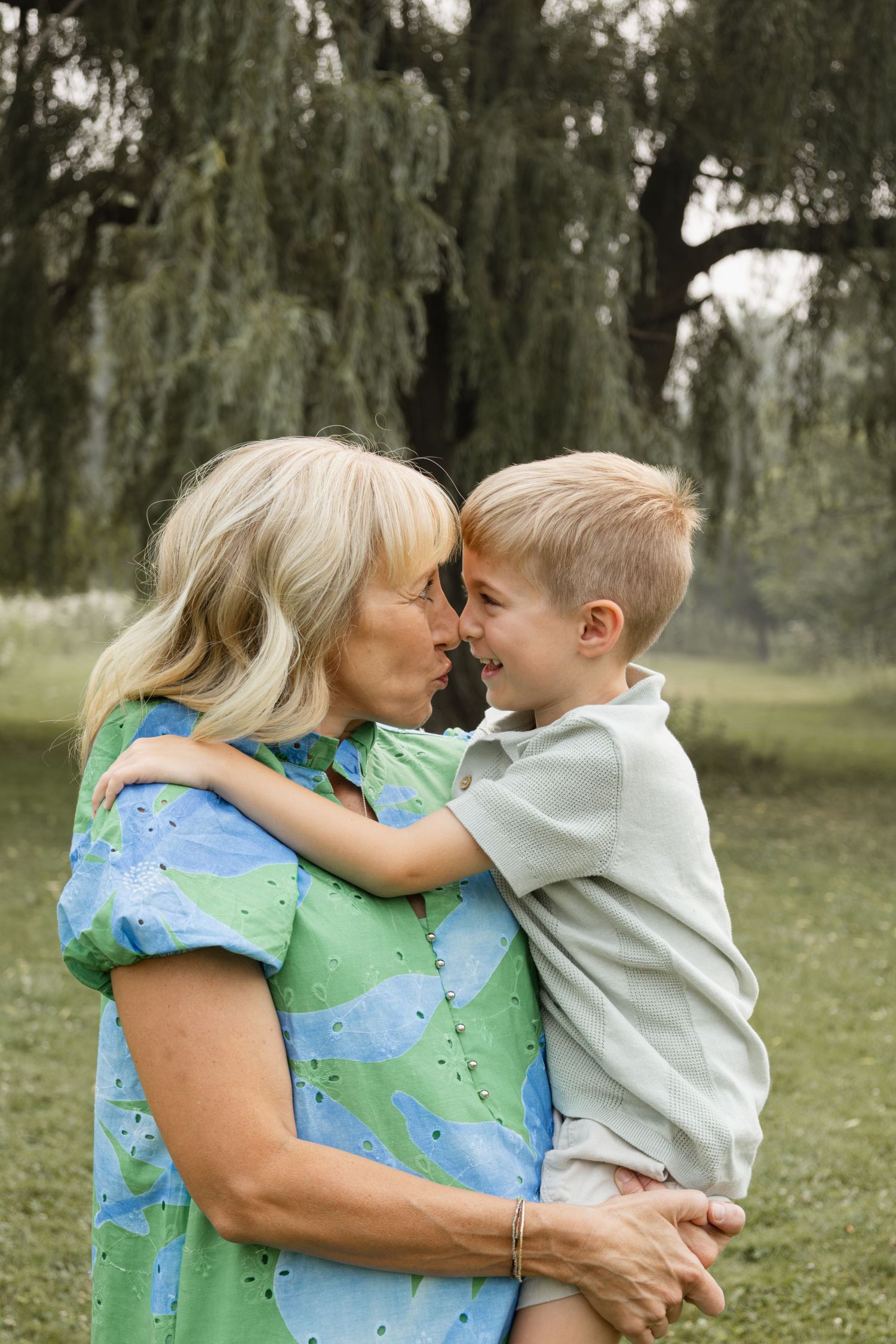 Grandmother and young boy share a tender nose-to-nose moment while embracing outdoors near a willow tree.