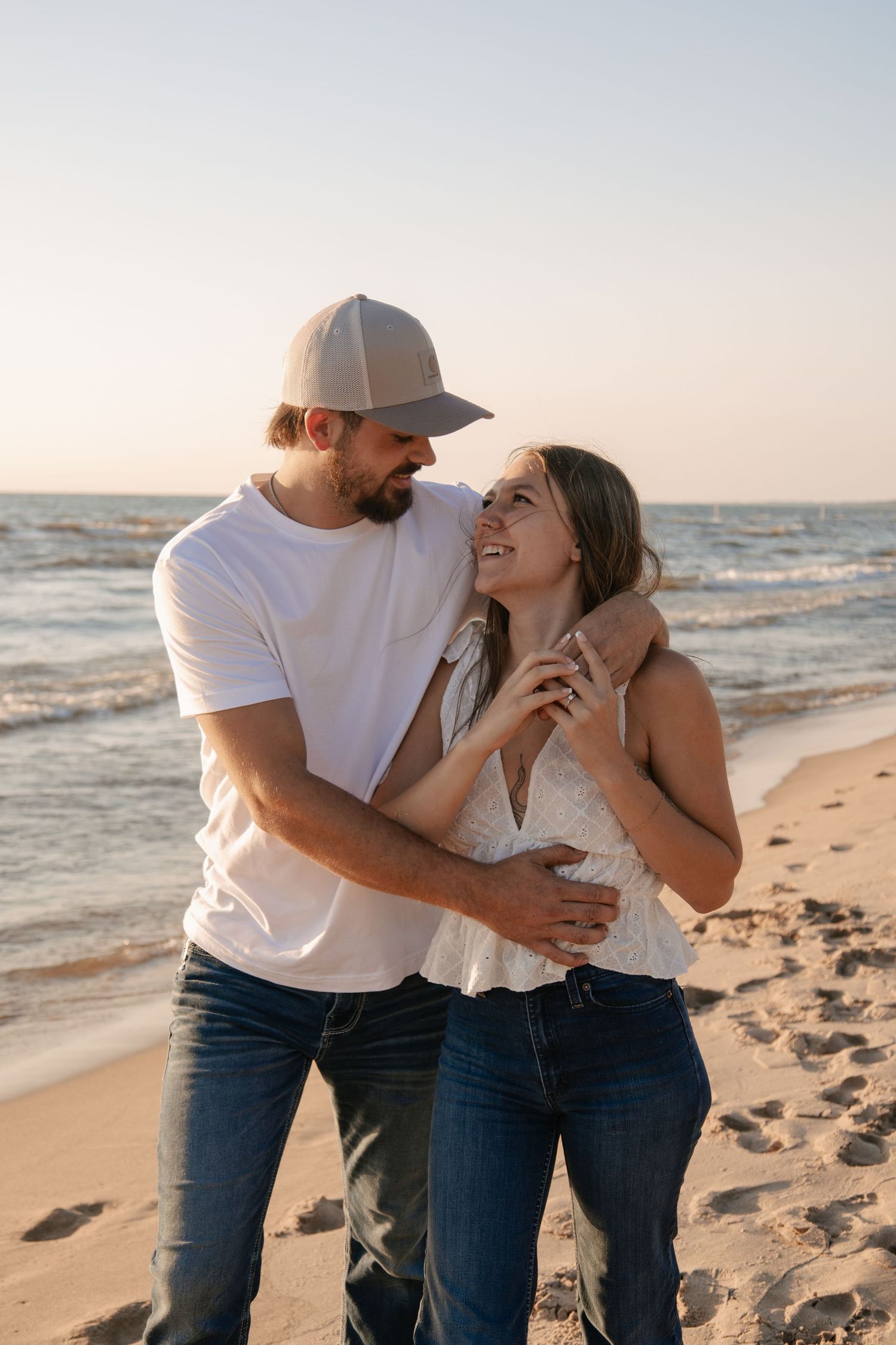 A couple embraces and smiles at each other on a sandy beach at sunset, man in white tee and cap, woman in white top.