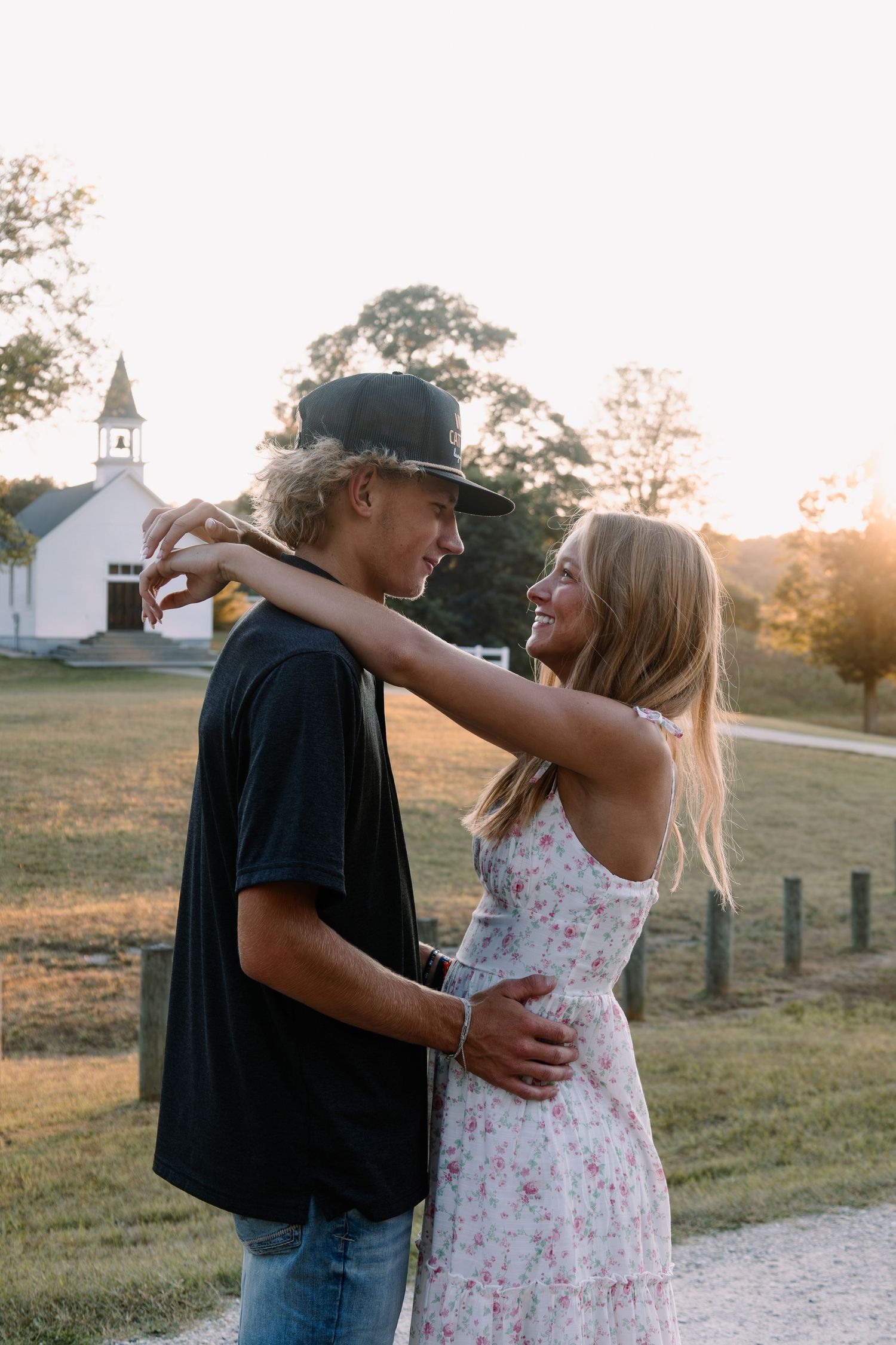 Young couple embracing at sunset near a white country church, smiling at each other in a romantic outdoor setting.