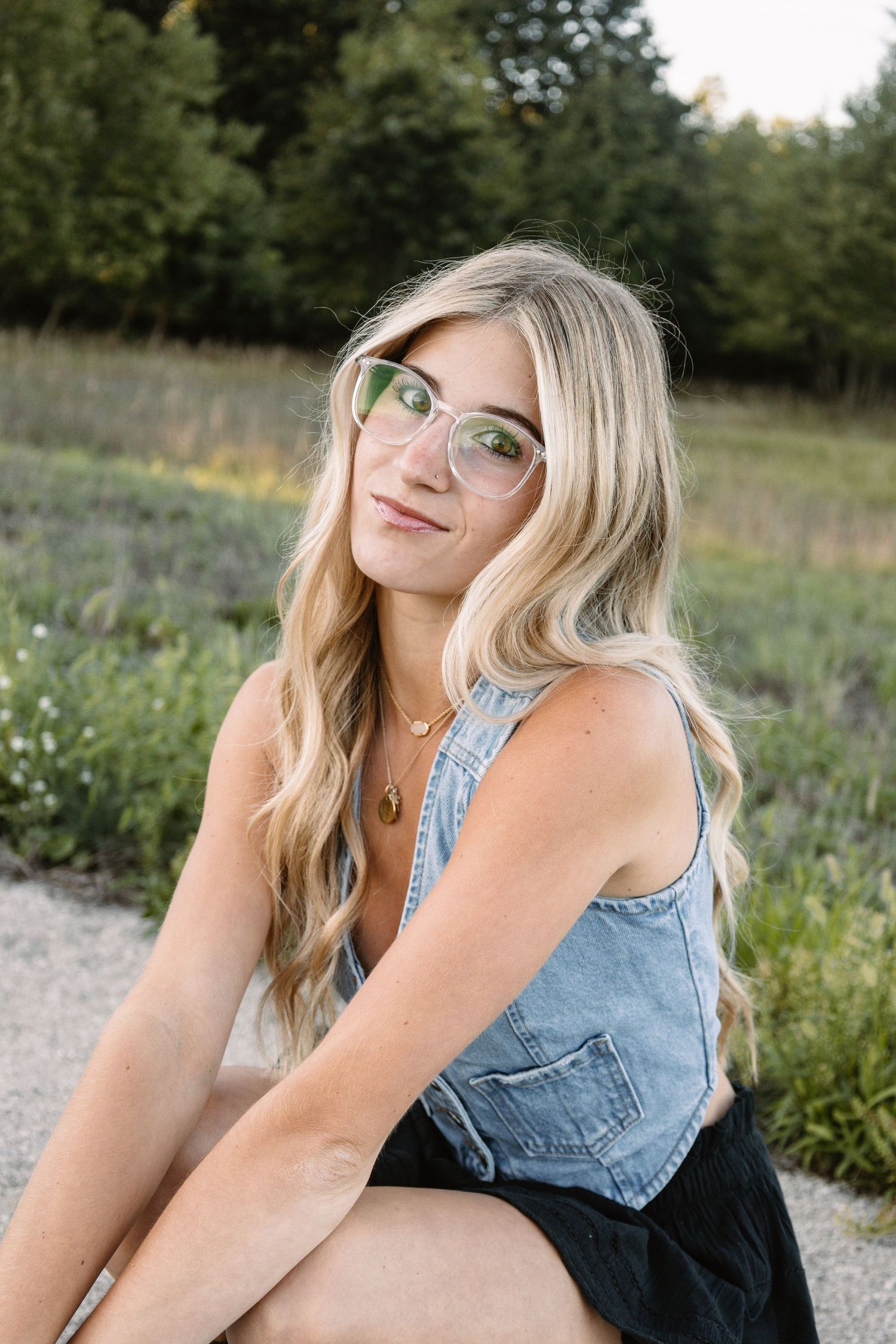 Blonde woman with clear glasses in denim vest sitting outdoors in field with wildflowers.