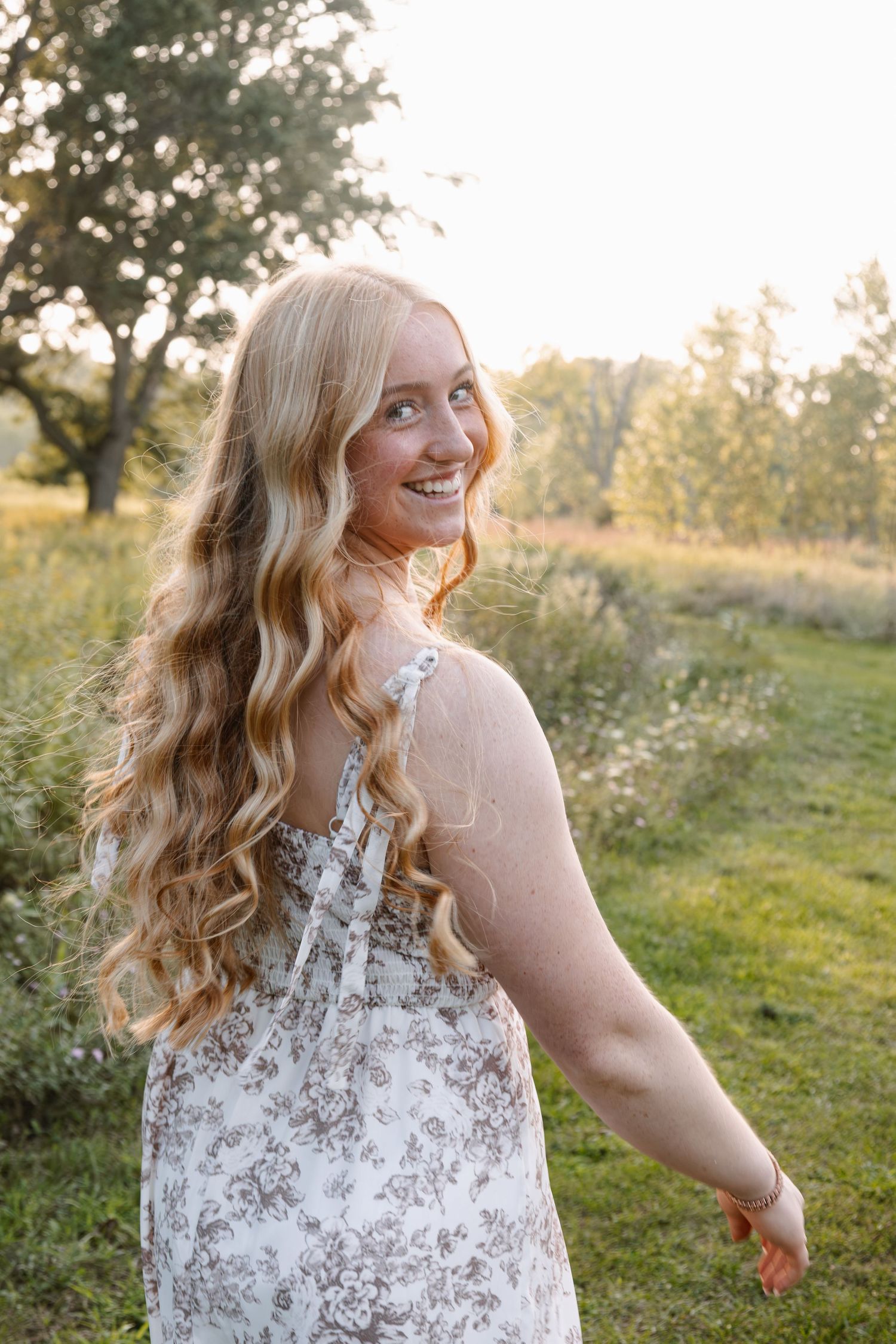 Young blonde woman in floral sundress smiling outdoors in golden hour sunlight with lush green trees behind her.