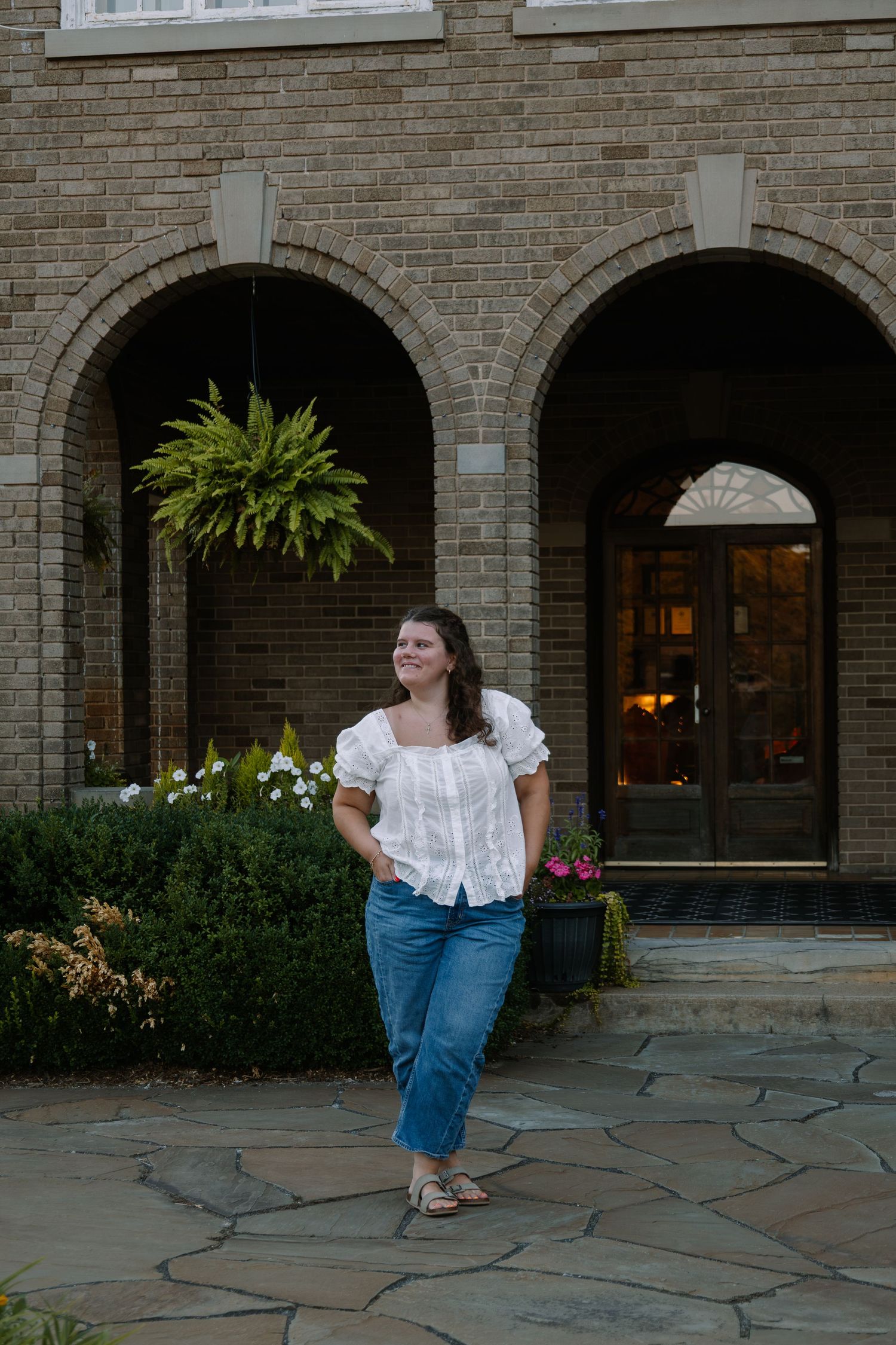 Woman in white top and jeans standing on stone patio outside brick building with arched doorways and lush greenery.