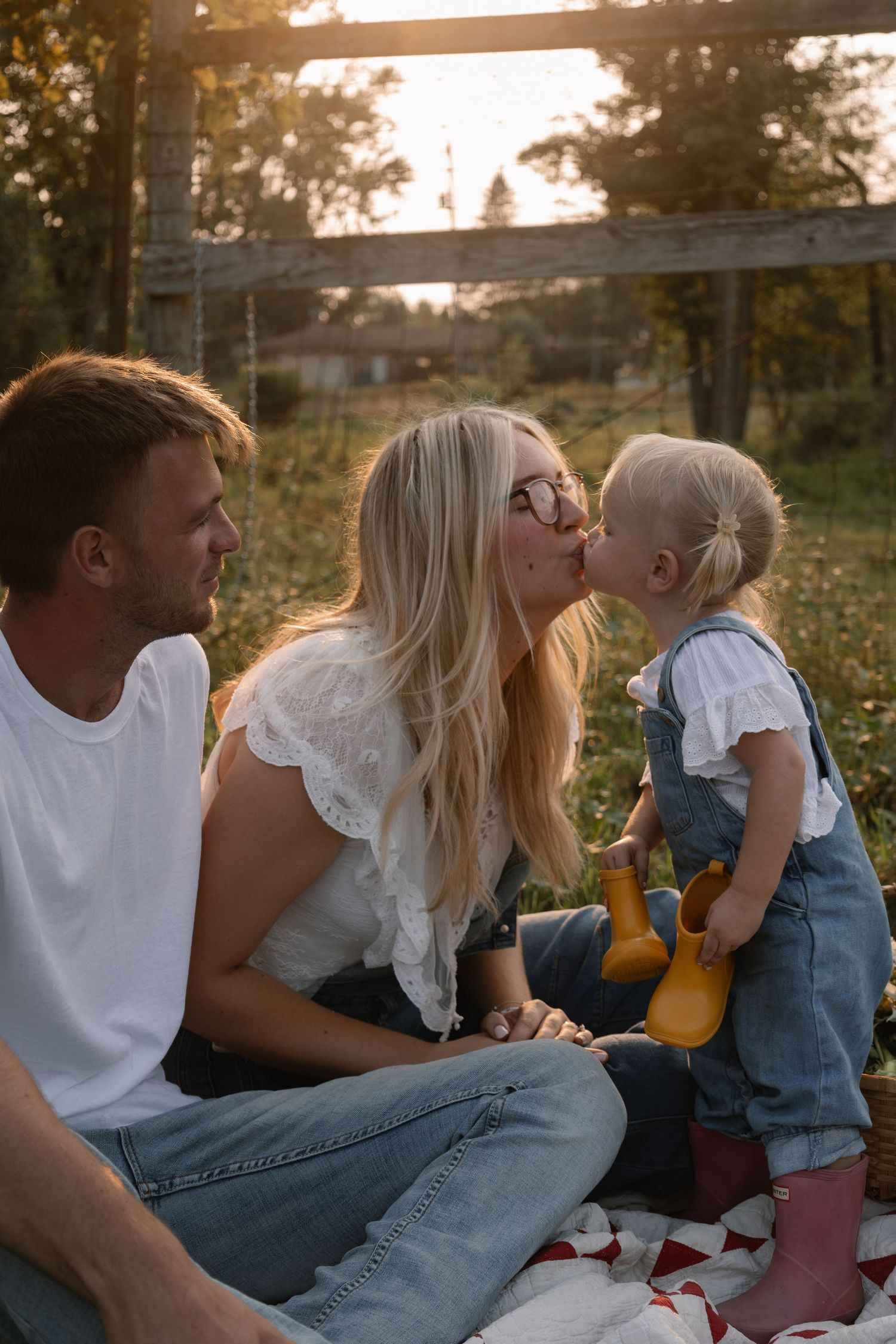 A mother shares a sweet kiss with her toddler daughter while father watches during a golden hour family picnic outdoors.