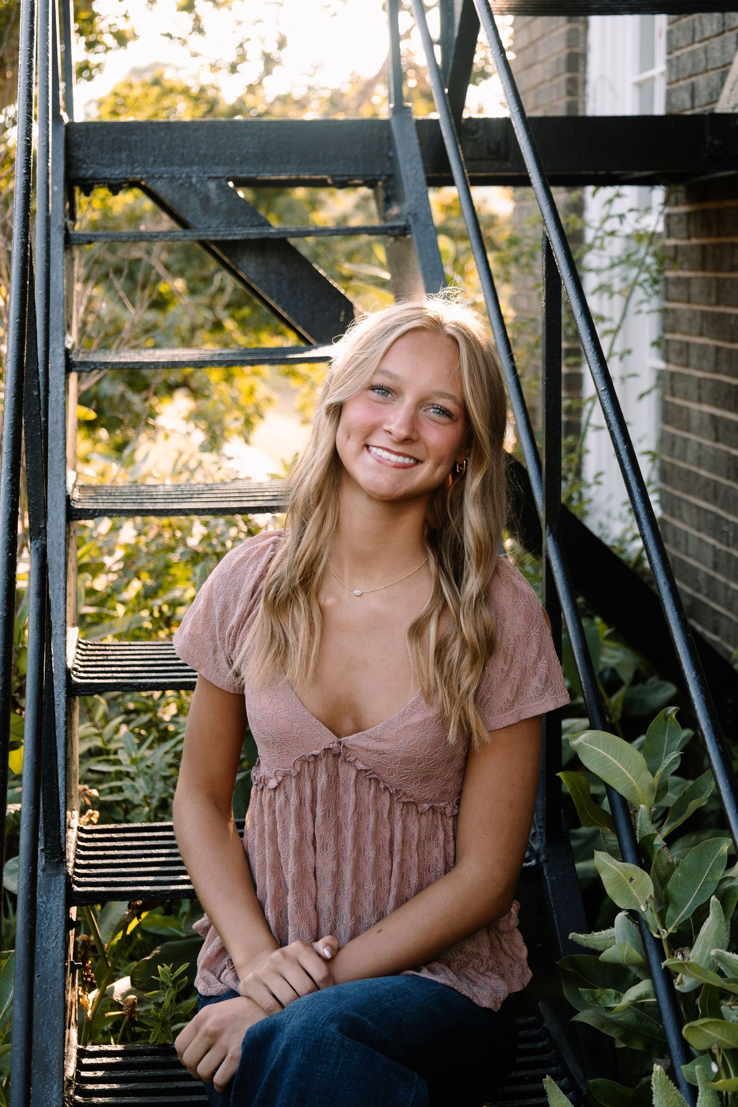 Smiling blonde teen girl sitting on metal spiral staircase surrounded by lush greenery in warm sunlight.