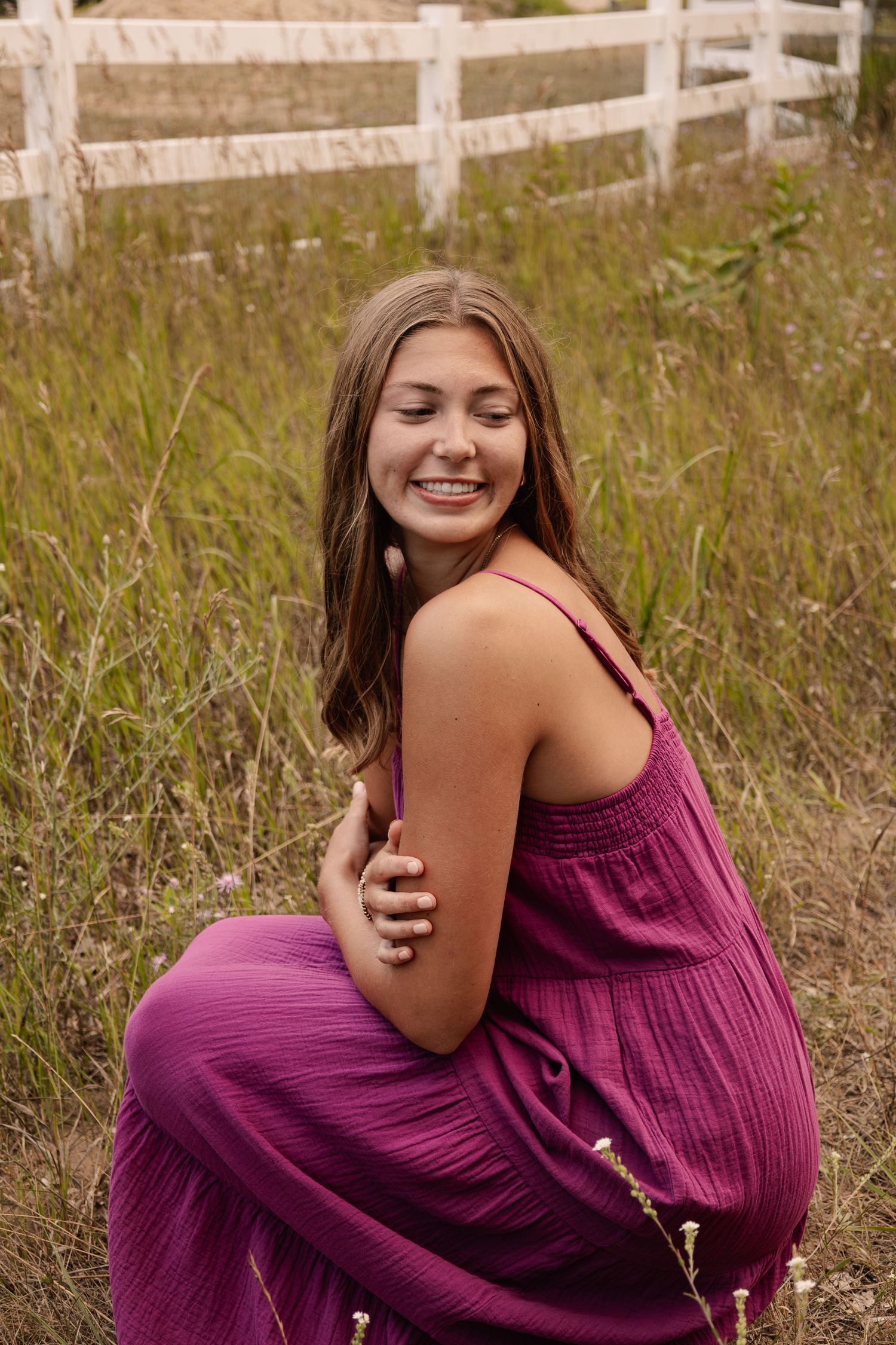 A young woman in a magenta jumpsuit sits smiling in a grassy field near a white wooden fence.