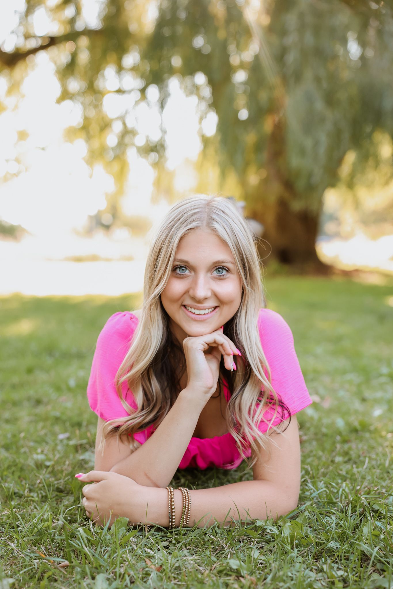Smiling blonde woman in pink dress lying on grass under a willow tree in golden sunlight.