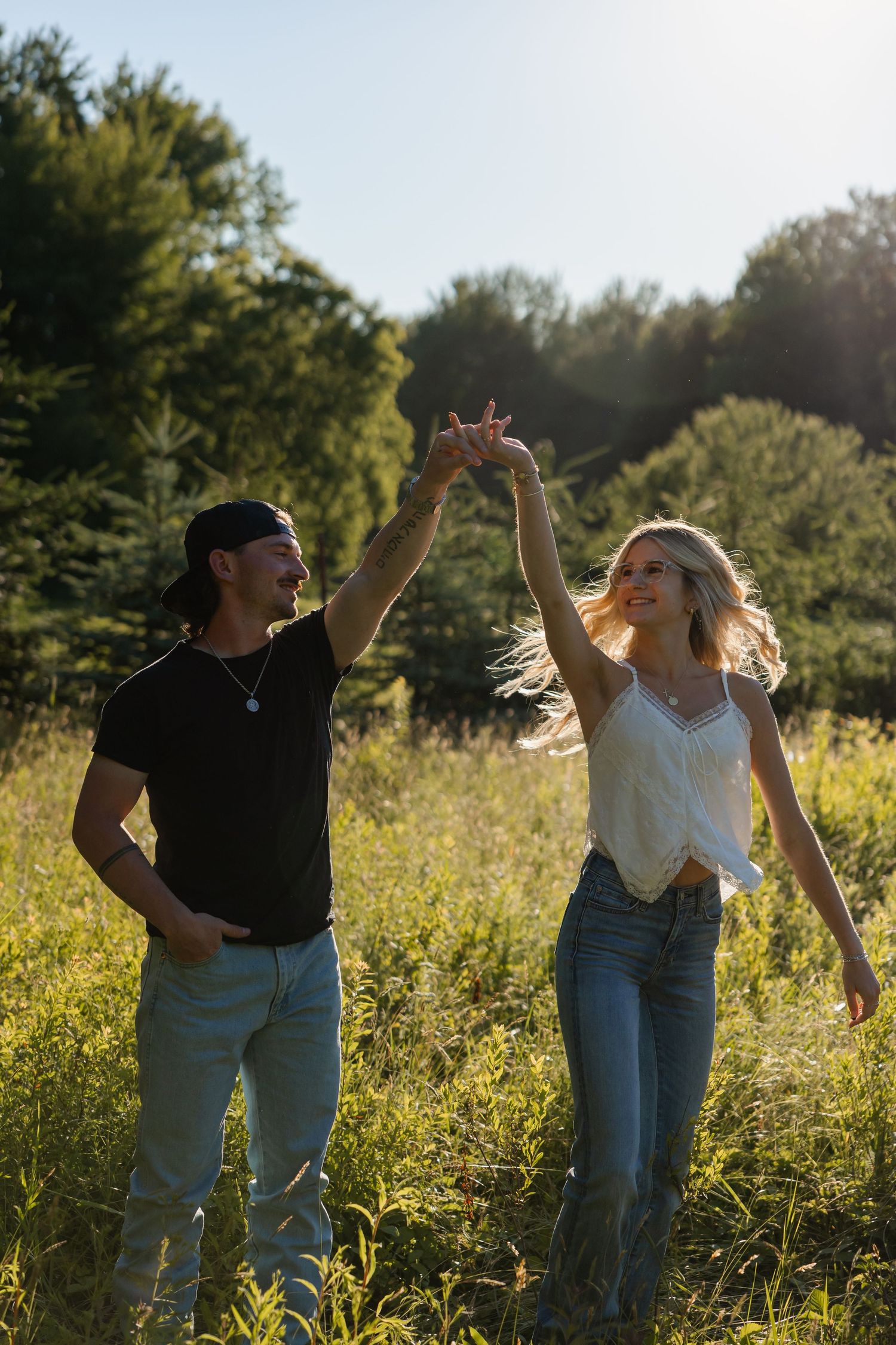 Joyful couple dancing in a sunny field, man spinning woman with raised hands, both wearing jeans and casual tops.