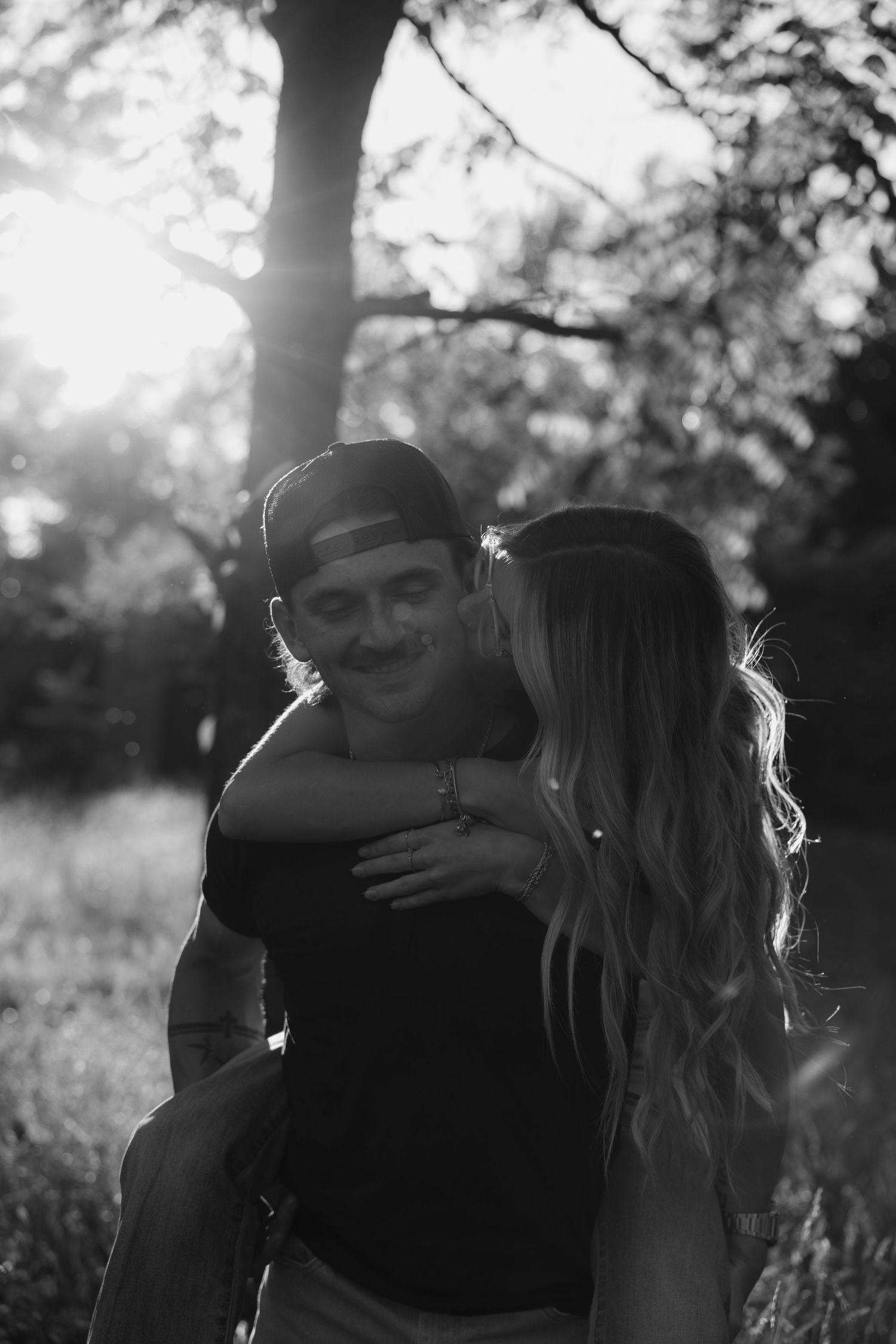 Black and white romantic photo of woman kissing mans cheek while he smiles, backlit by golden sunlight through trees.