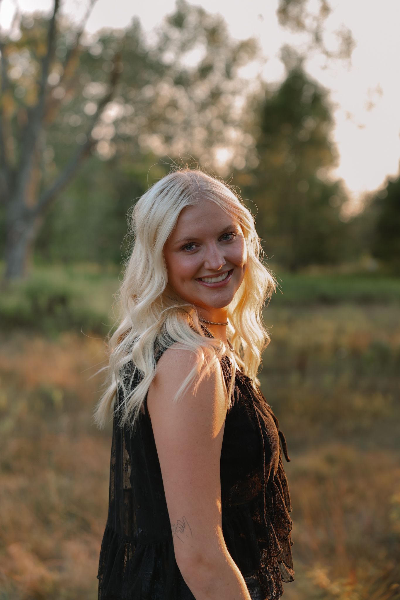 Smiling blonde woman in black fringe dress with choker necklace posing outdoors in a field during golden hour sunset.