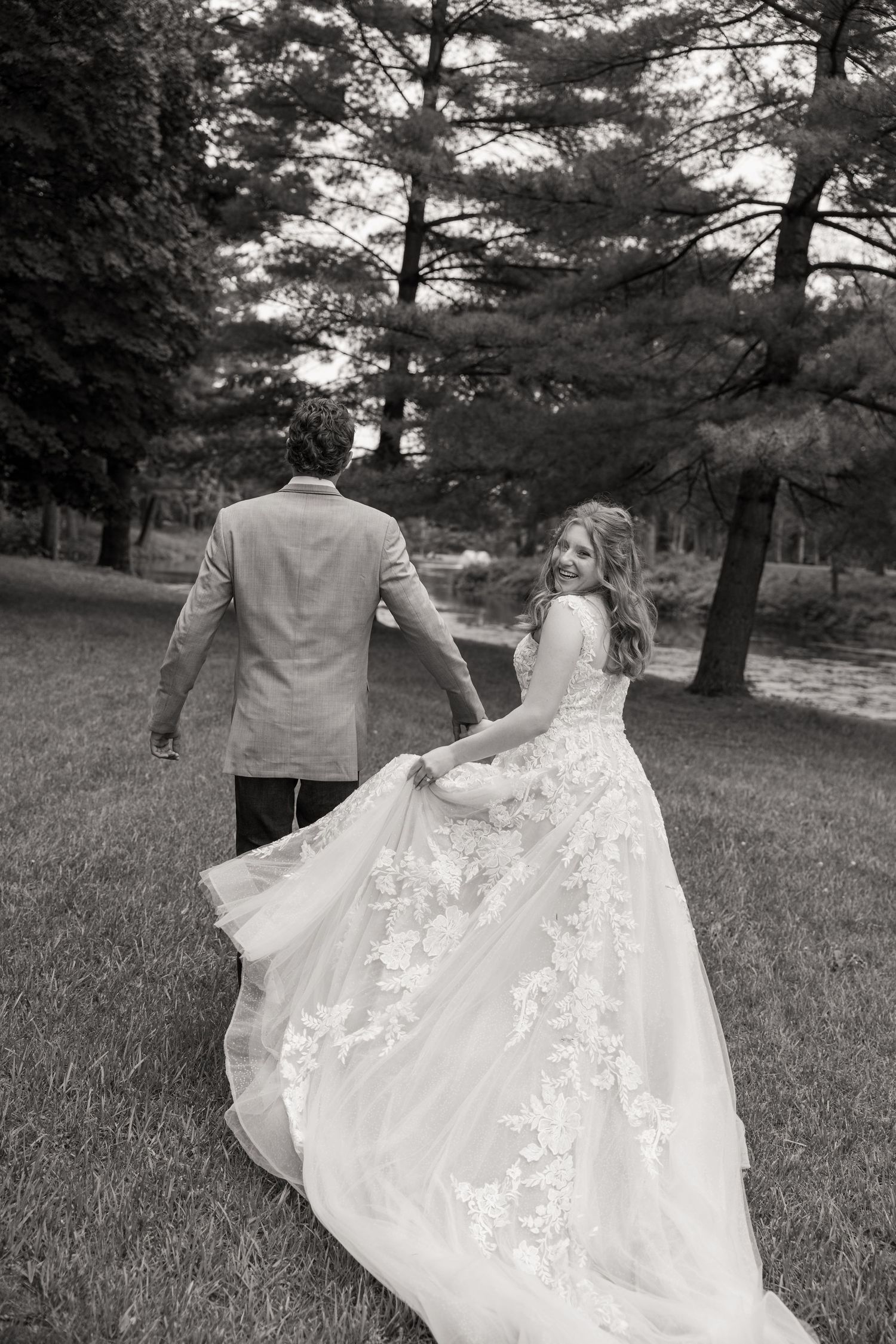 Black and white photo of bride and groom walking hand in hand through a green park.