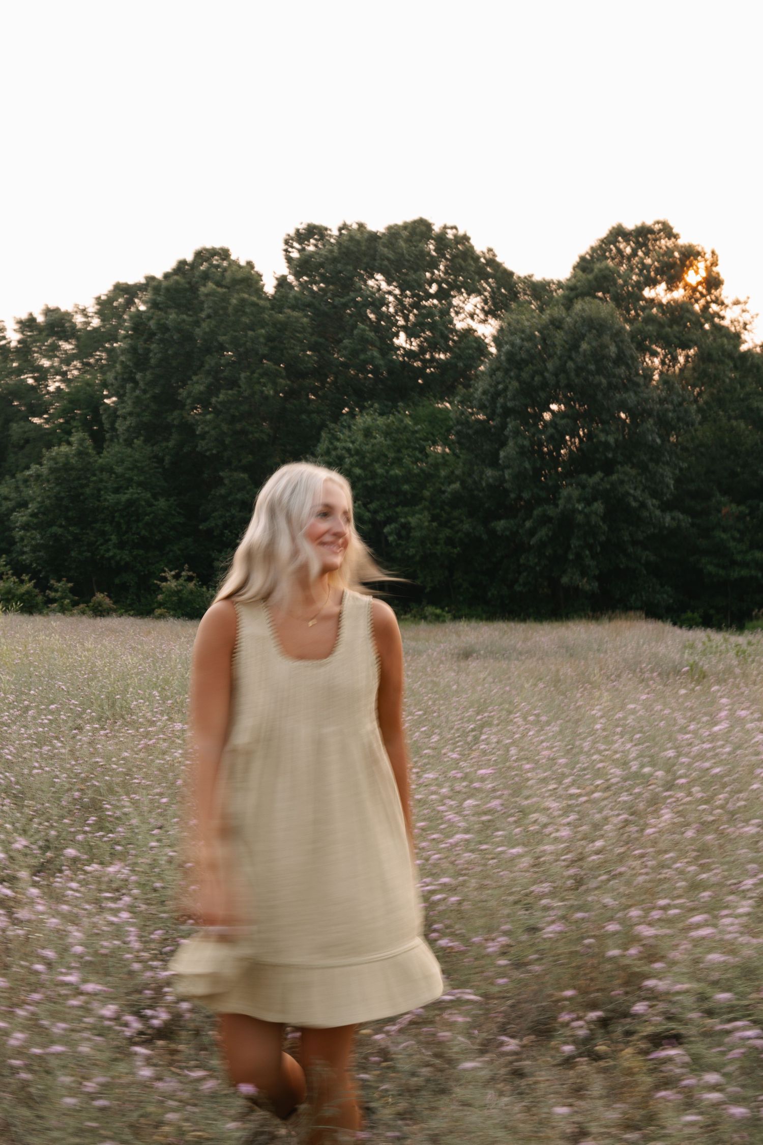 A smiling blonde woman in a beige linen dress twirls in a wildflower meadow at dusk.