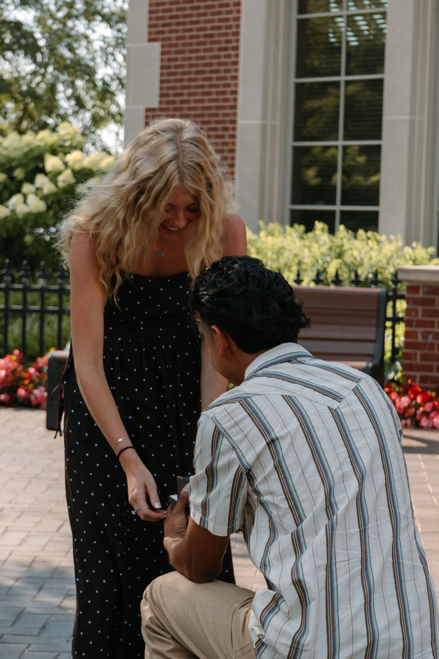 Man kneeling on one knee proposing to a smiling blonde woman in a black polka dot dress outdoors.