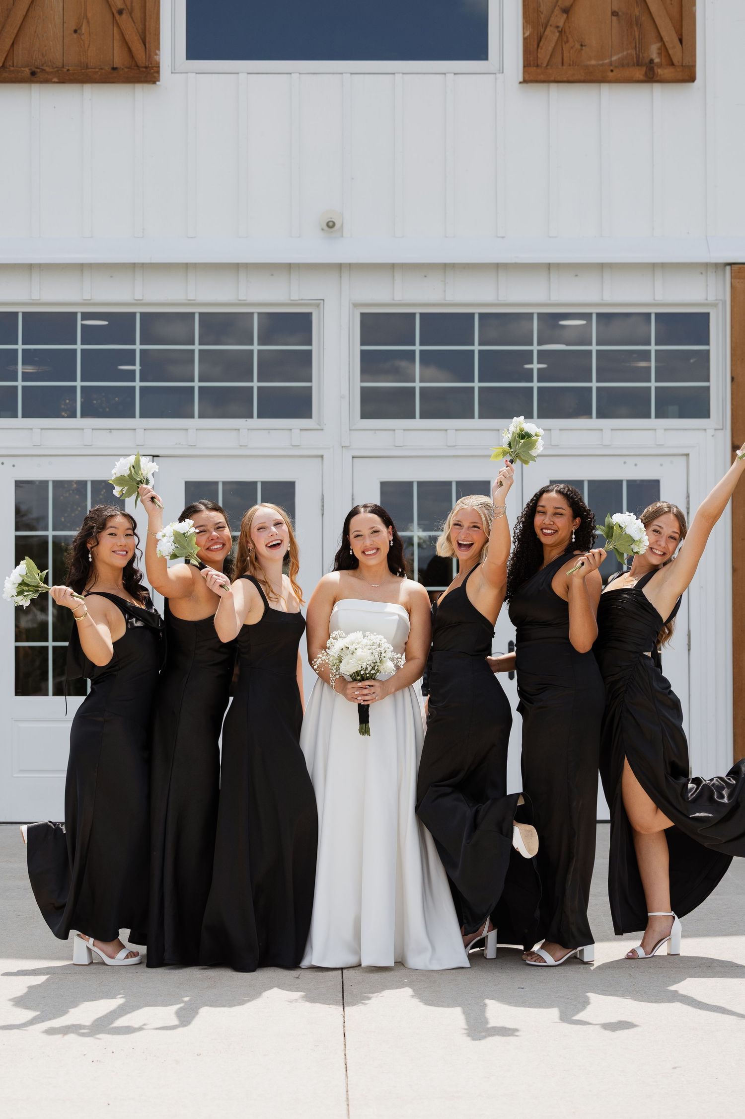 Bride in white gown with six bridesmaids in black dresses celebrating joyfully outside a white barn wedding venue.