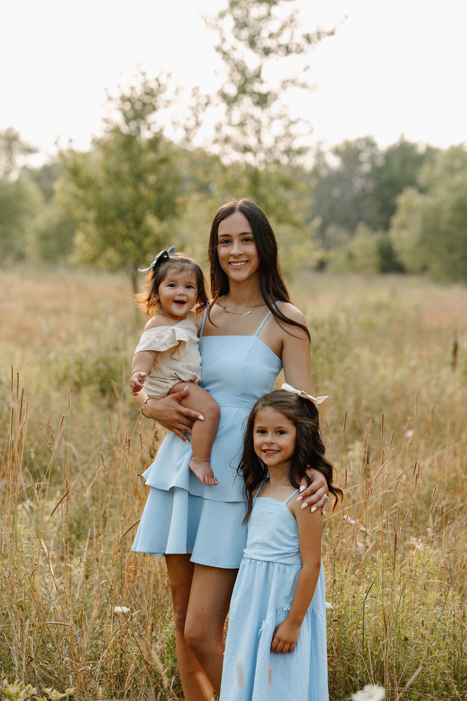 A smiling woman in a blue dress holds a baby while a young girl in matching blue stands beside her in a sunny field.