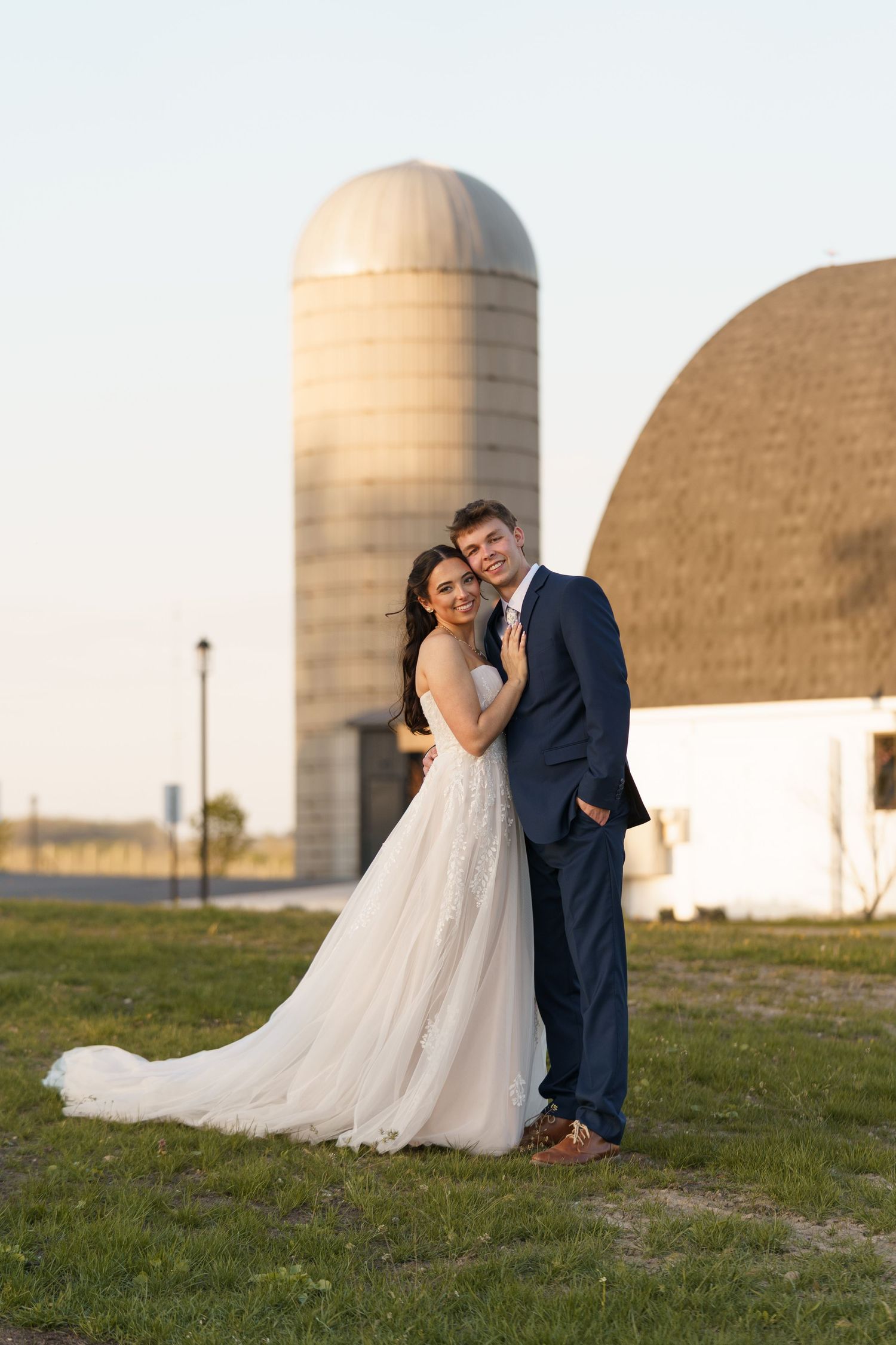 A smiling bride and groom pose together in front of a rustic barn and silo.