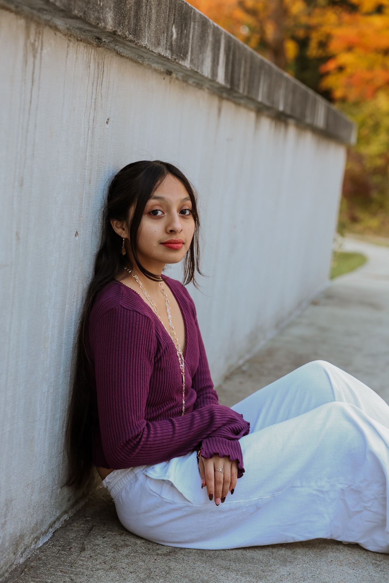 Young woman in burgundy ribbed top and white pants leaning against concrete wall in autumn.
