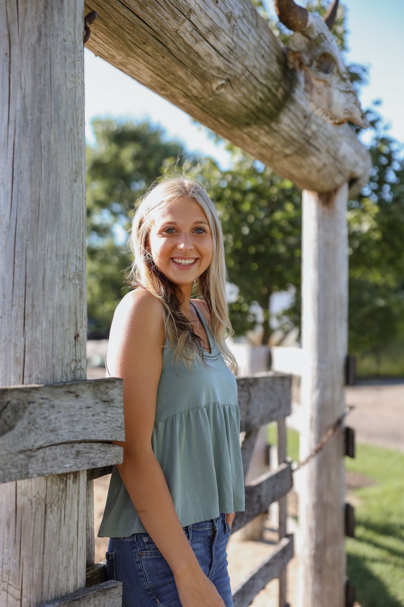 Smiling blonde woman in sage green tank top leaning against a rustic wooden fence outdoors.