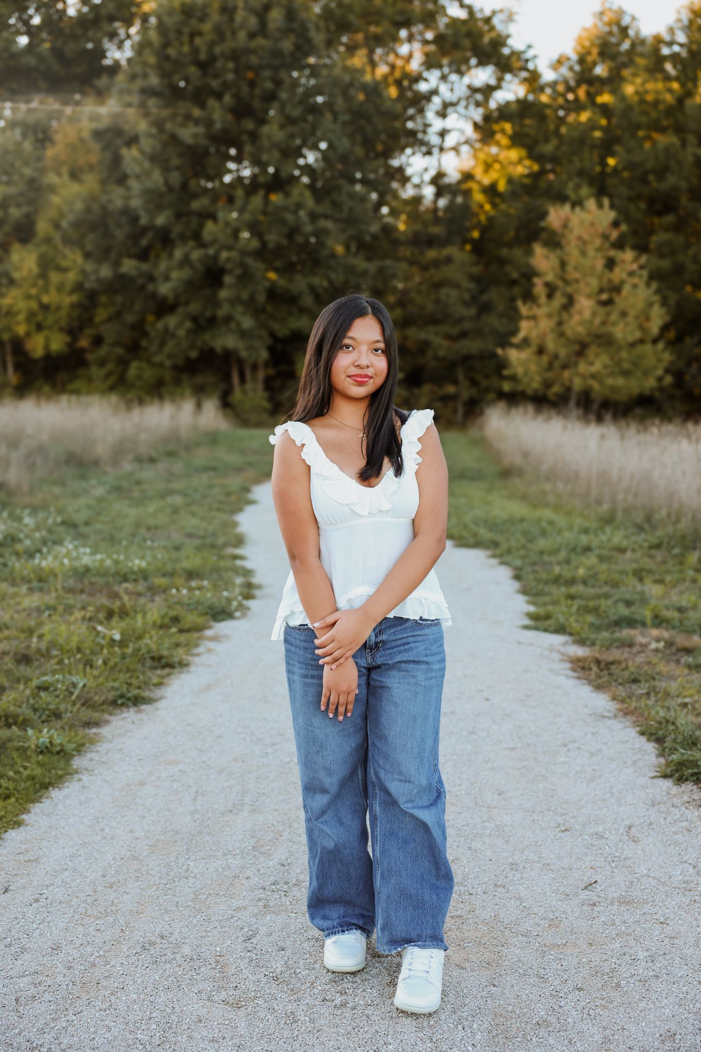 Young Asian woman in white ruffled top and wide-leg jeans stands on a scenic gravel park path.