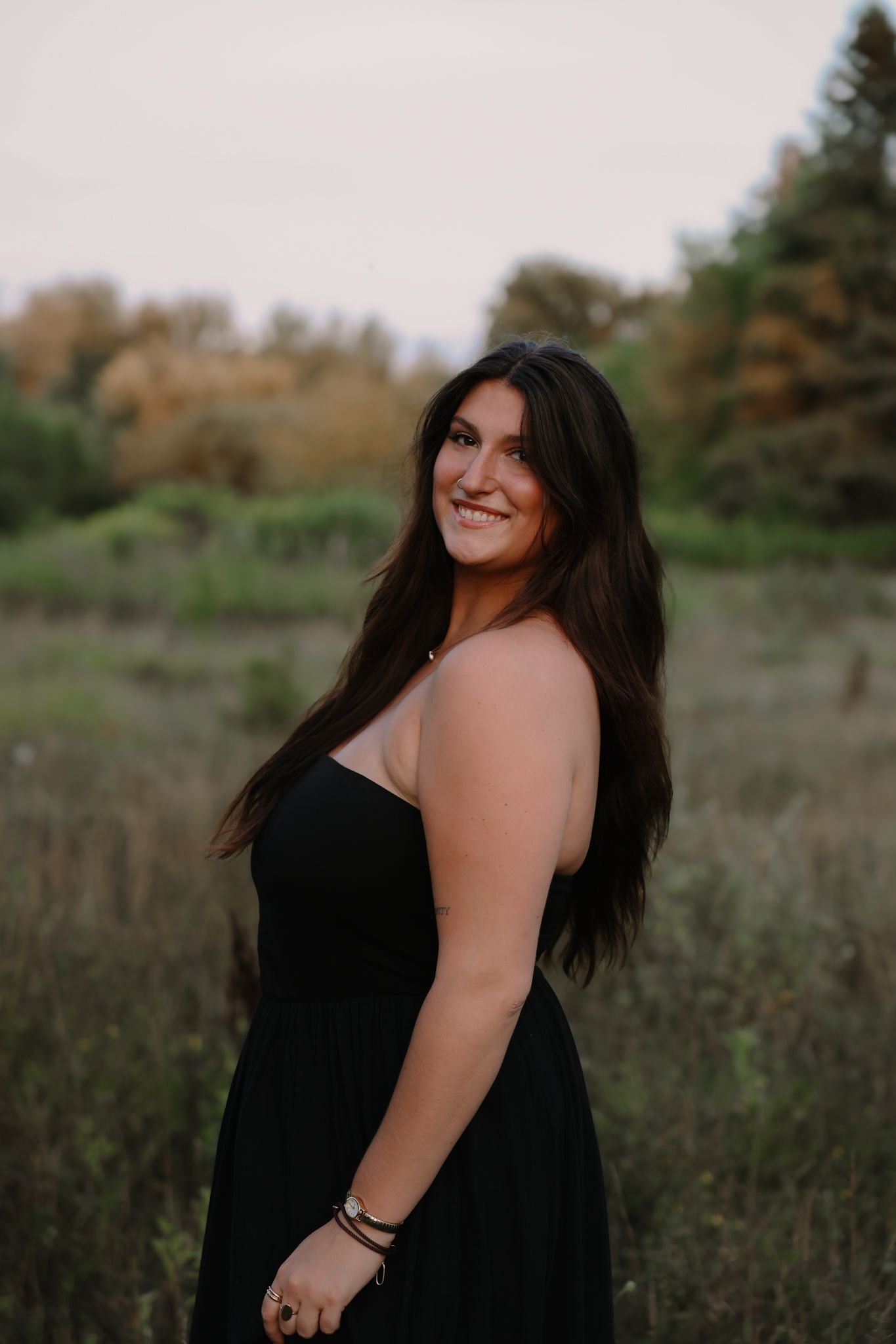 Smiling brunette woman in black strapless dress posing in a green field during a soft overcast outdoor photo session.