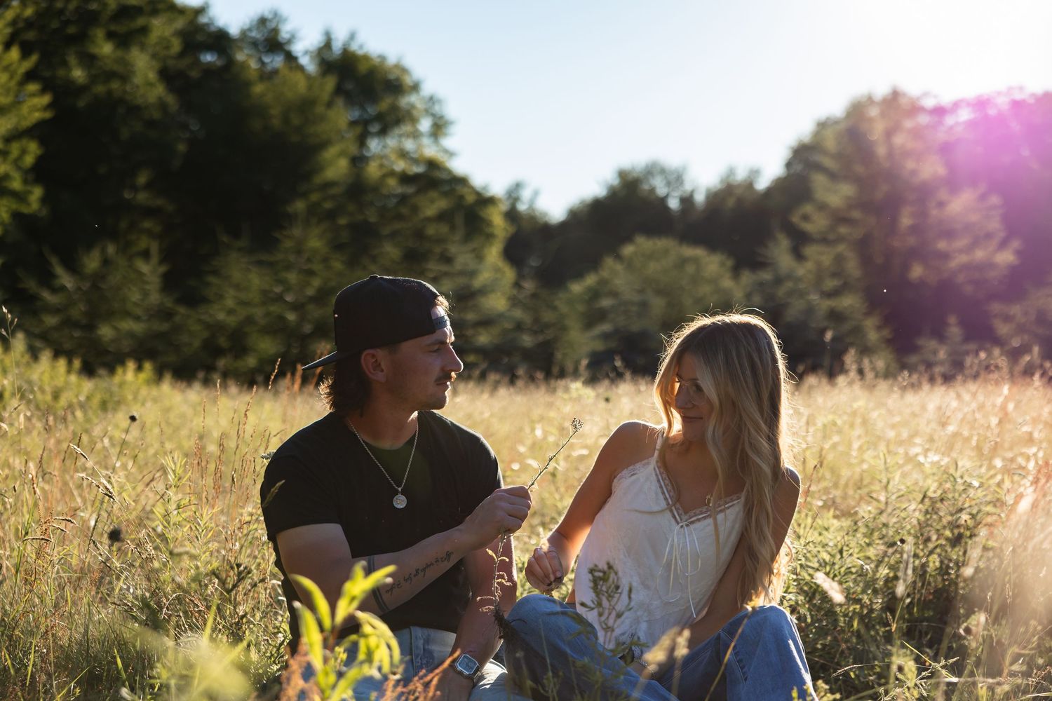 Couple sitting together in a golden sunlit meadow, gazing at each other surrounded by tall grass and trees.