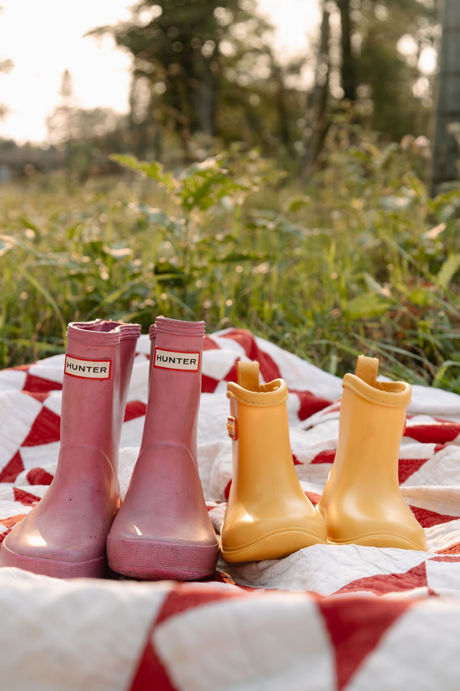 Two pairs of Hunter rain boots, pink and yellow, displayed on a red and white quilt in a natural outdoor setting.