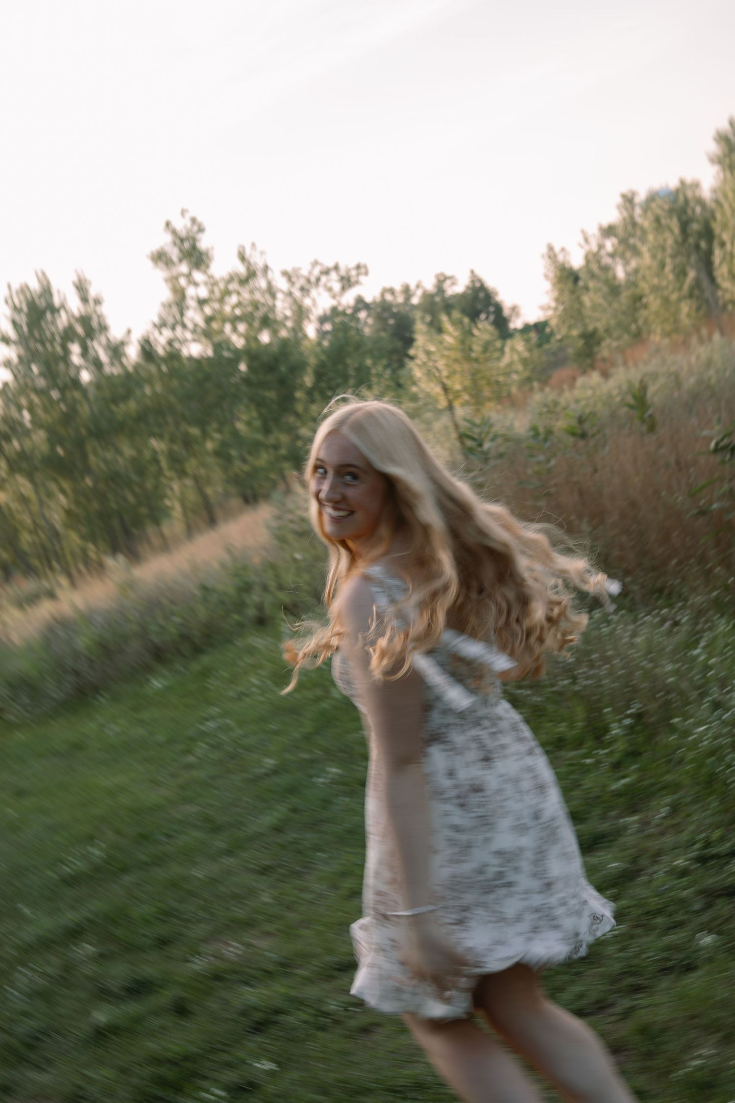 Blonde woman in floral dress spinning joyfully in a green meadow at golden hour.