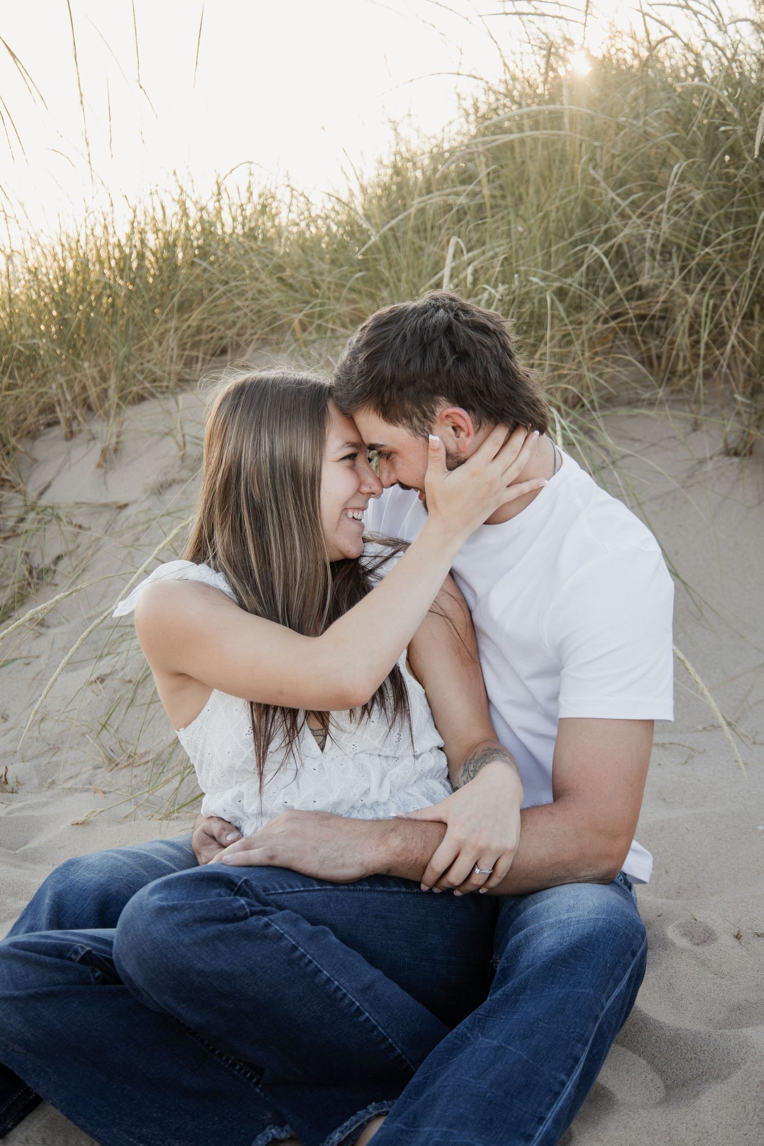Couple sitting on sandy beach dunes, smiling and embracing romantically, surrounded by coastal grass.